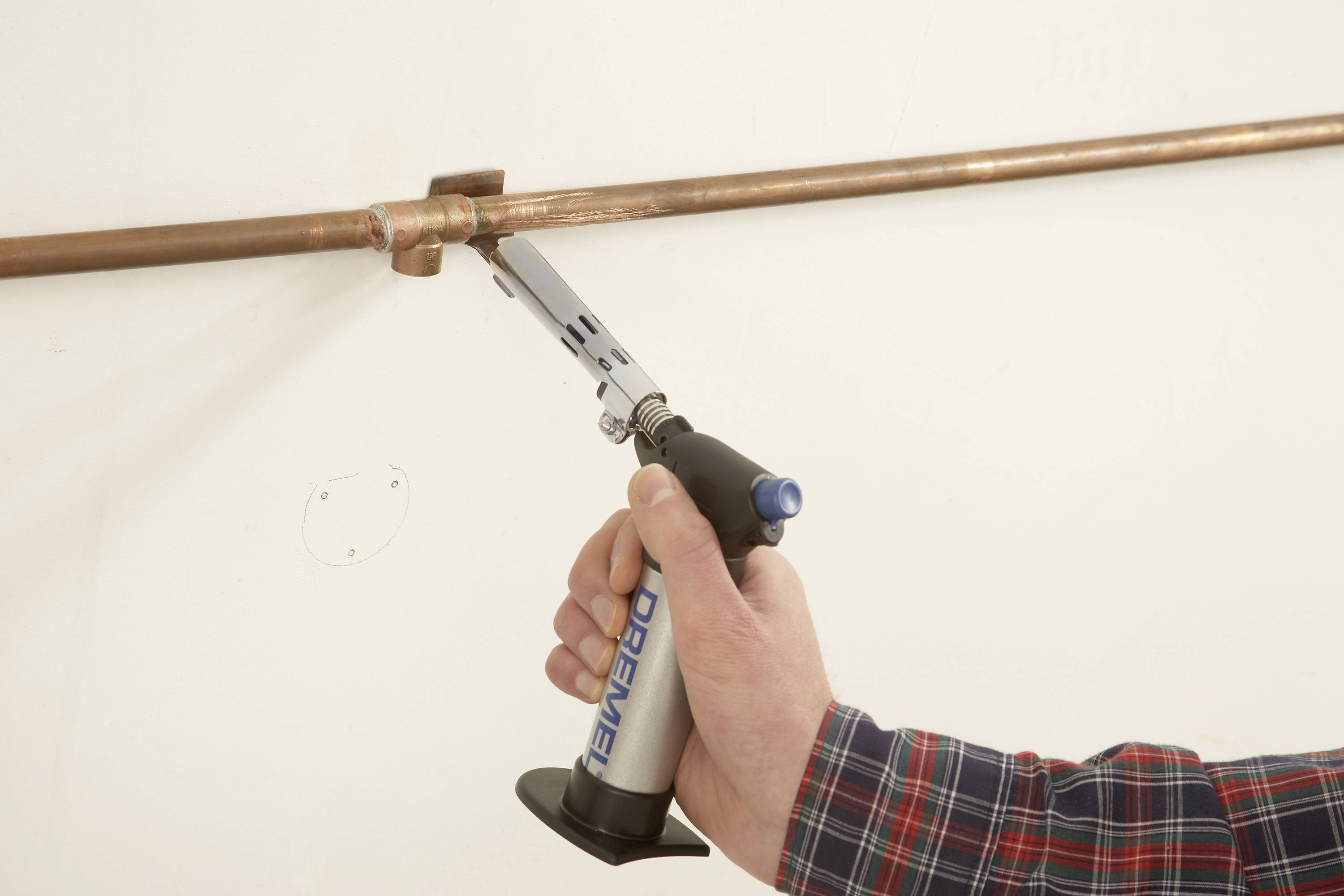 Eine Person lötet ein Kupferrohr mit einem Handbrenner an einer weißen Wand. Der Brenner liegt in der rechten Hand.