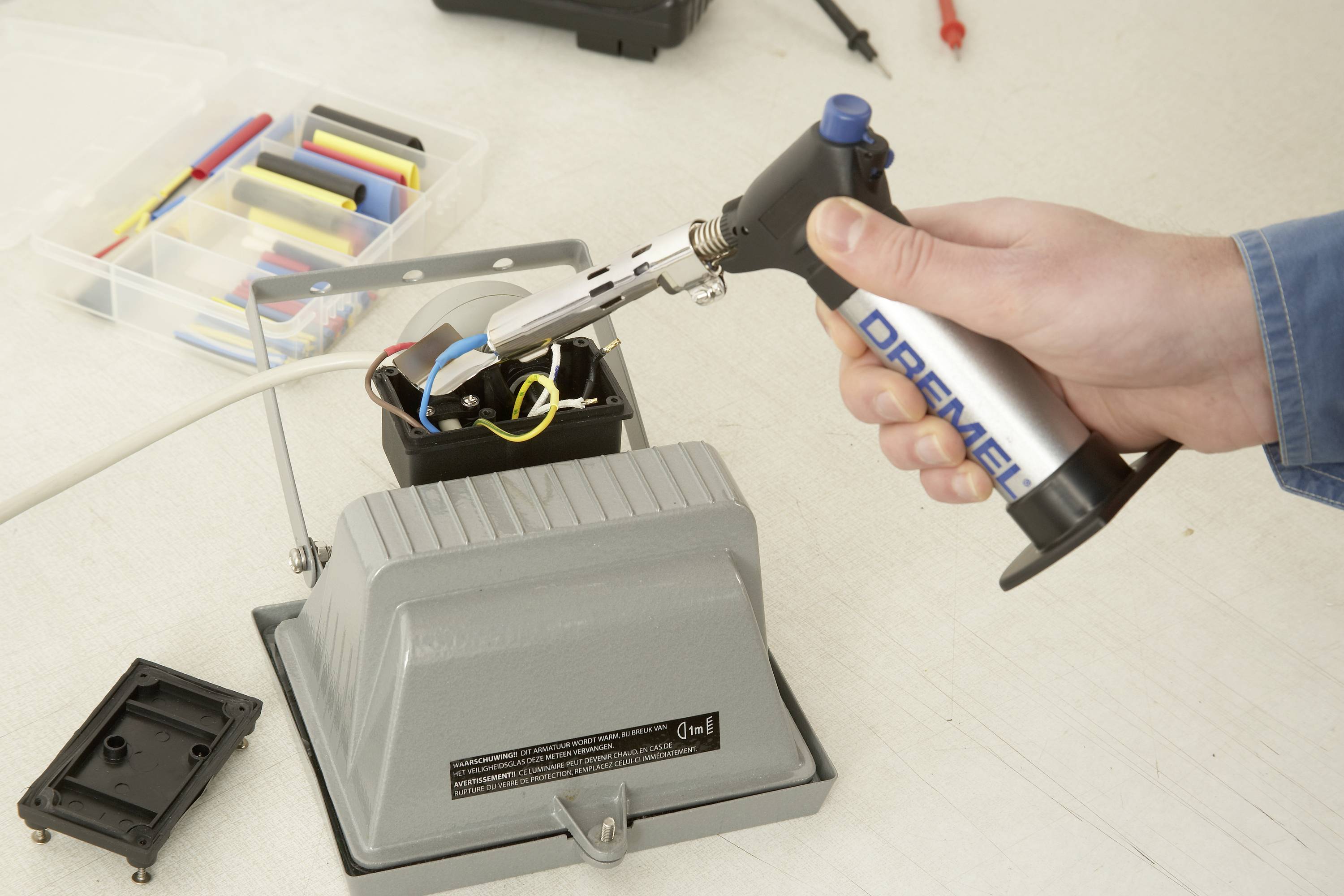 Eine Person verwendet einen Handbrenner, um an einer elektrischen Lampe Drähte zu verbinden. Im Hintergrund ist Werkzeug zu sehen.