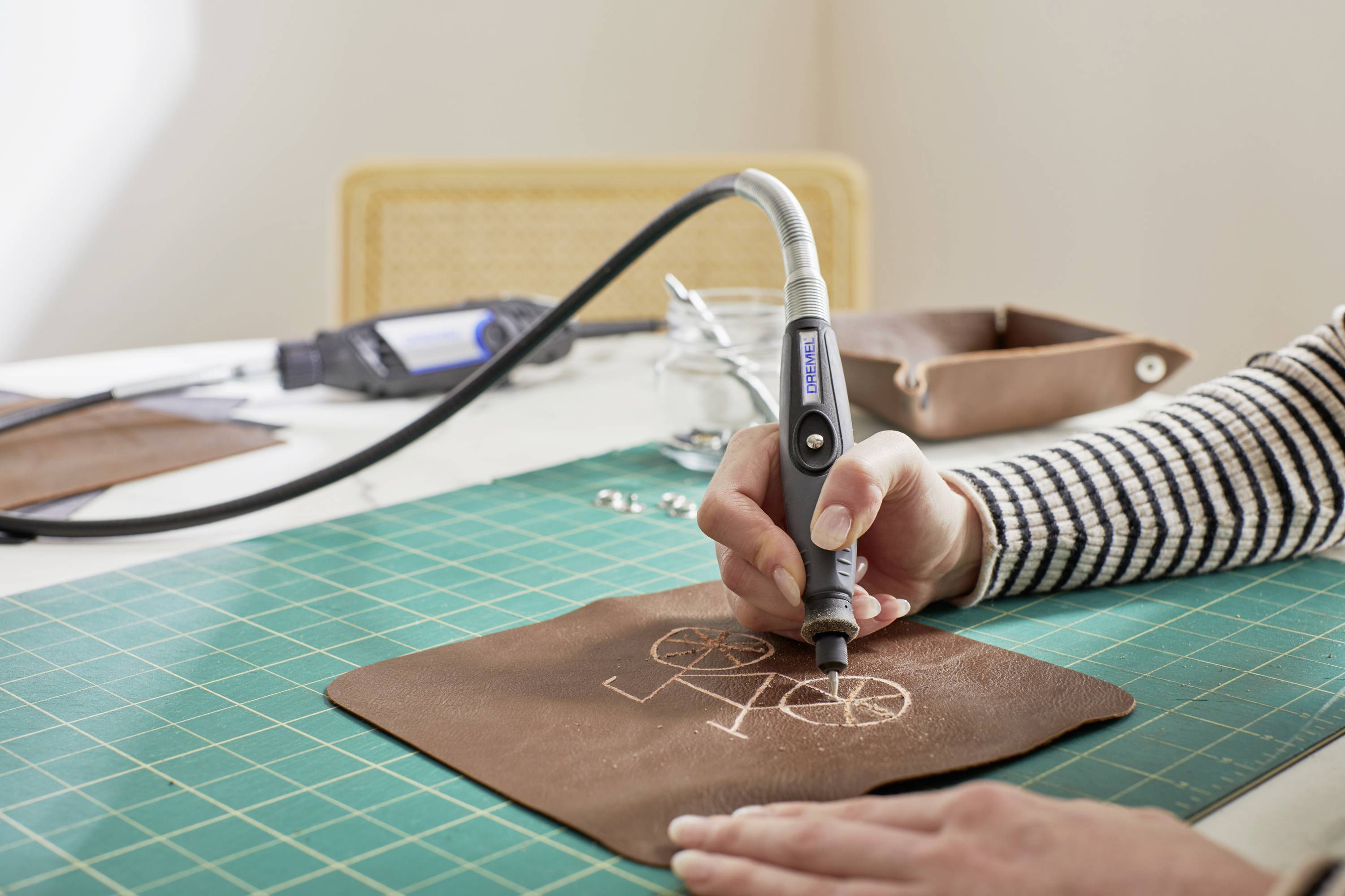 Eine Person graviert ein Fahrradmotiv in ein Stück braunes Leder auf einer grünen Schneidematte mit einem elektrischen Gravierstift.
