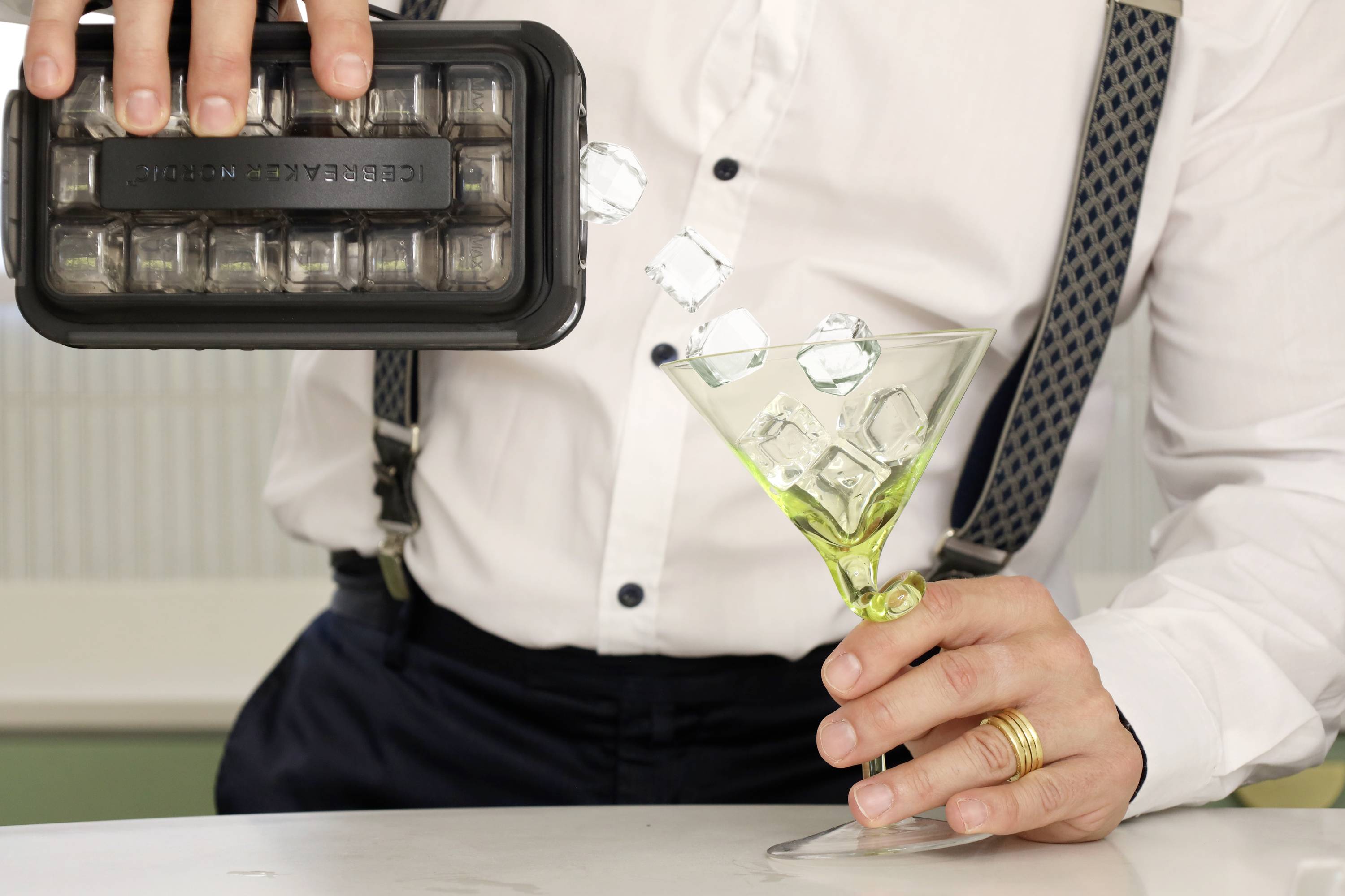 Eine Person mit weißen Hemd und Hosenträgern kippt Eiswürfel aus einer schwarzen Form in ein grünes Martini-Glas.