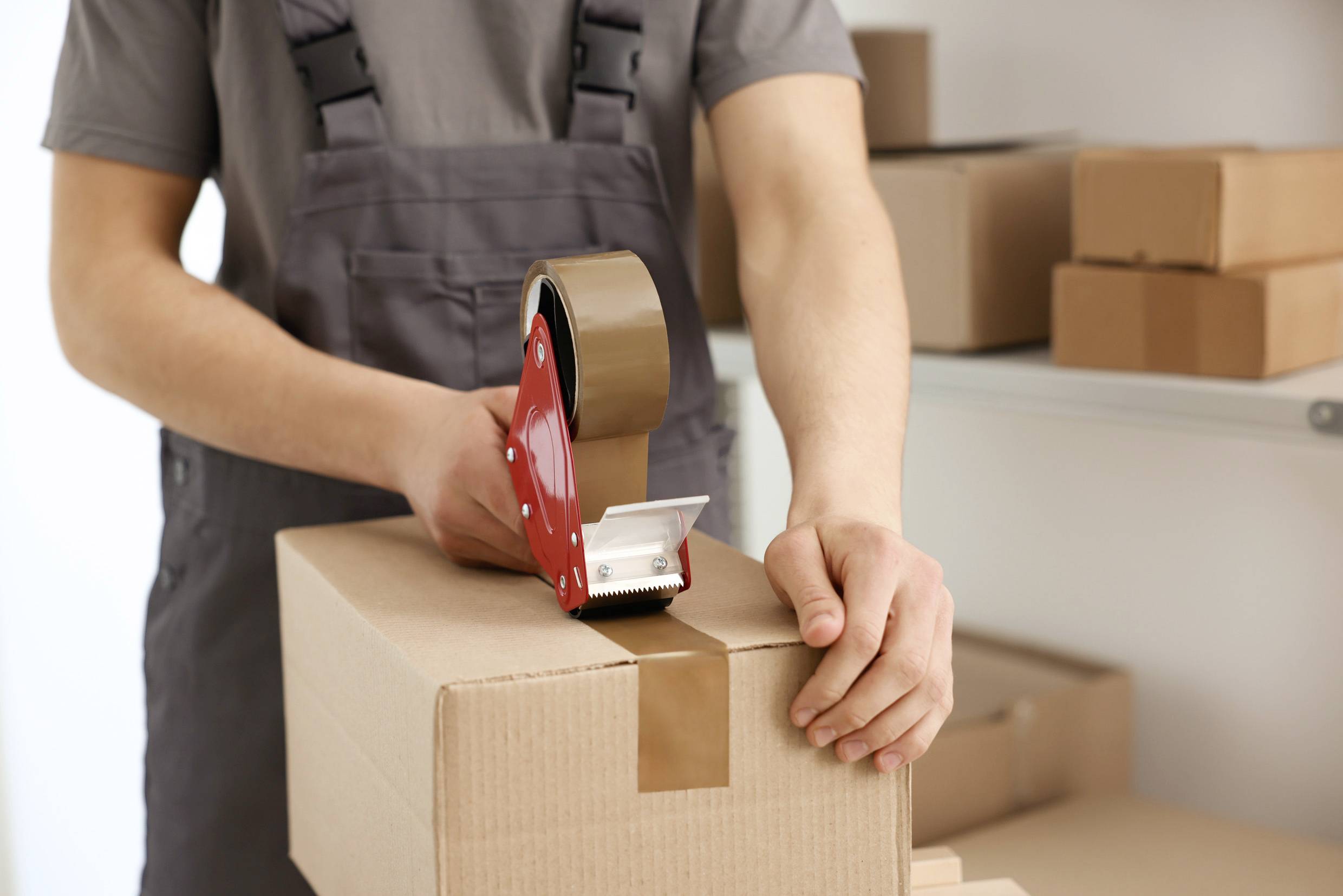 Eine Person verpackt einen Karton mit einem Klebebandabroller. Im Hintergrund sind weitere Kartons auf einem Regal zu sehen.