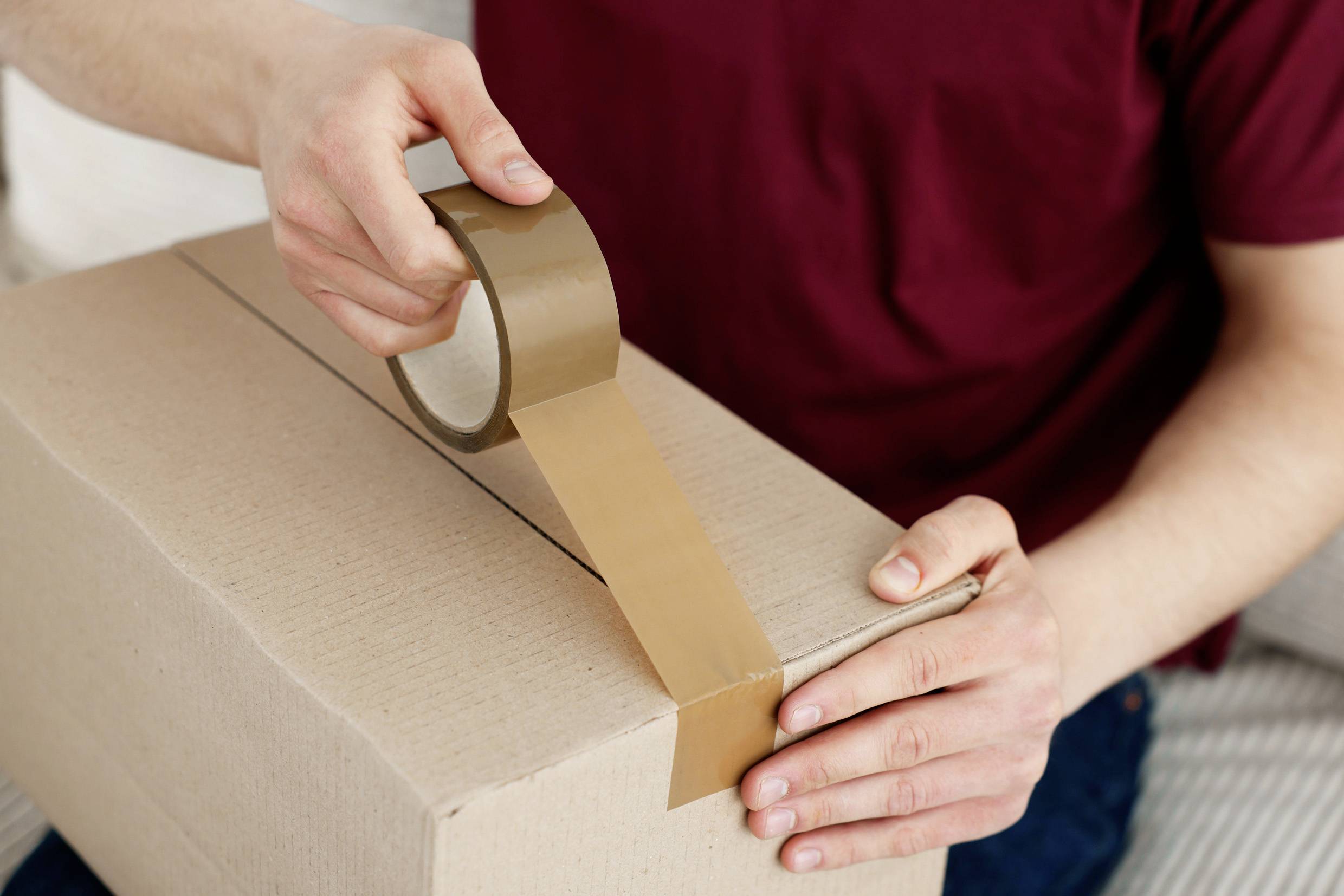 Eine Person klebt mit braunem Klebeband einen Karton zu. Der Karton wird vorbereitet, möglicherweise für Versand oder Umzug.
