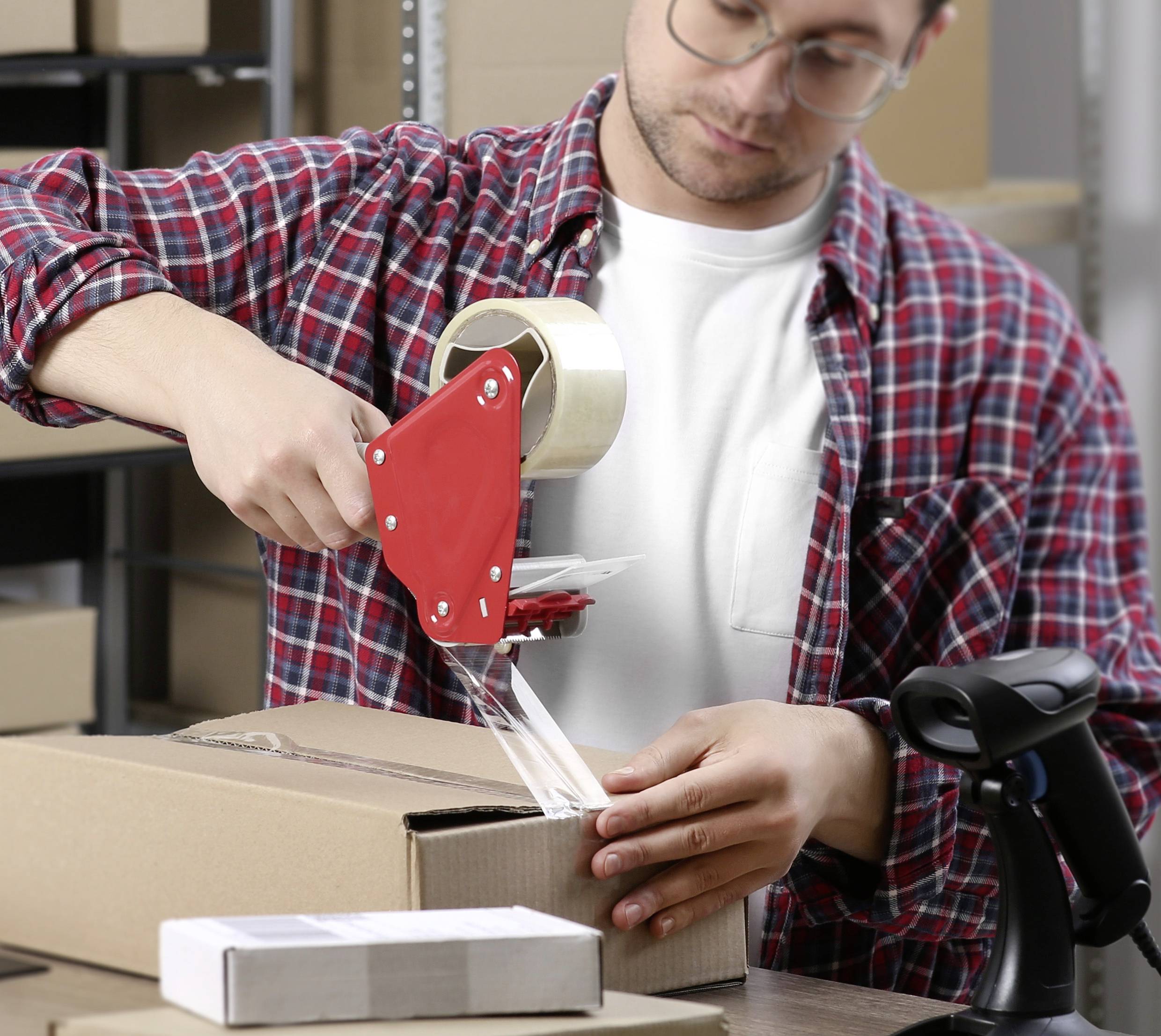 Ein Mann im karierten Hemd verschließt einen Karton mit Klebeband in einem Lager. Im Hintergrund sind Regale mit weiteren Kartons zu sehen.