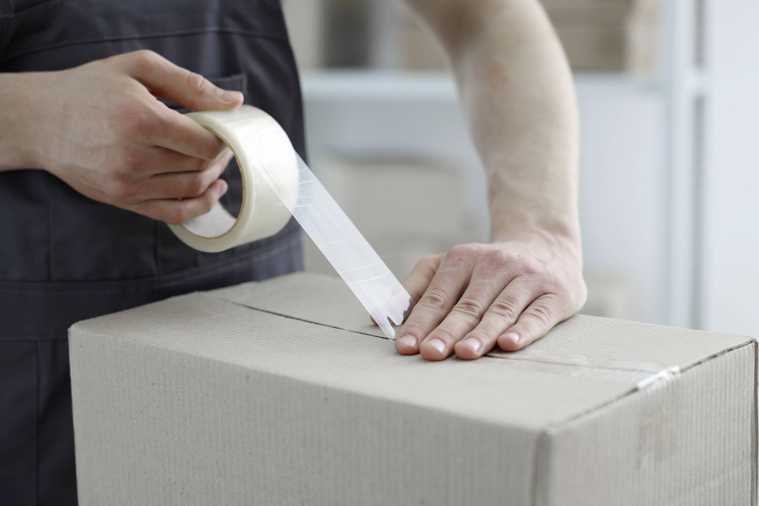 Eine Person klebt einen Karton mit transparentem Klebeband zu. Hände halten das Band und den Karton in einer Lagerumgebung.