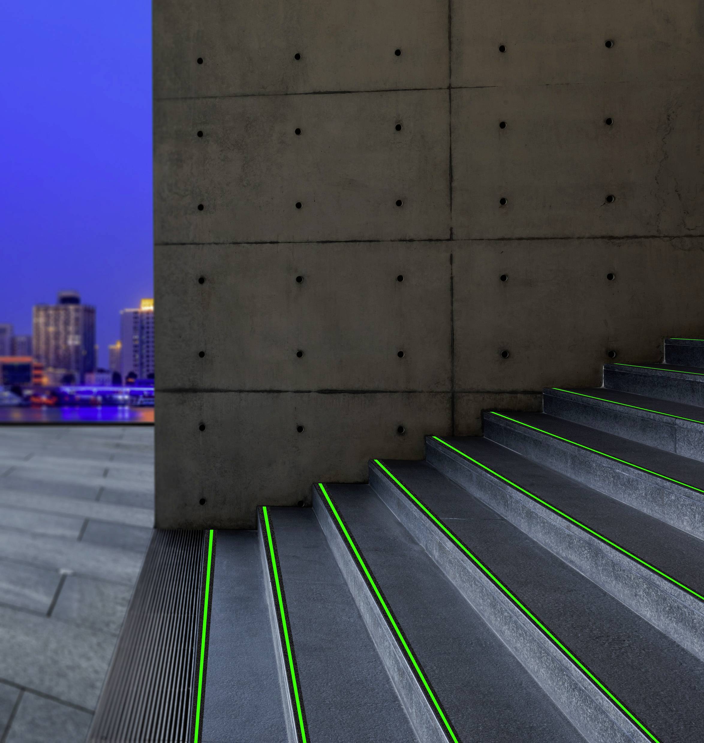 Eine moderne Treppe mit grün beleuchteten Stufenkanten neben einer Betonwand, im Hintergrund eine Stadtansicht bei Dämmerung.