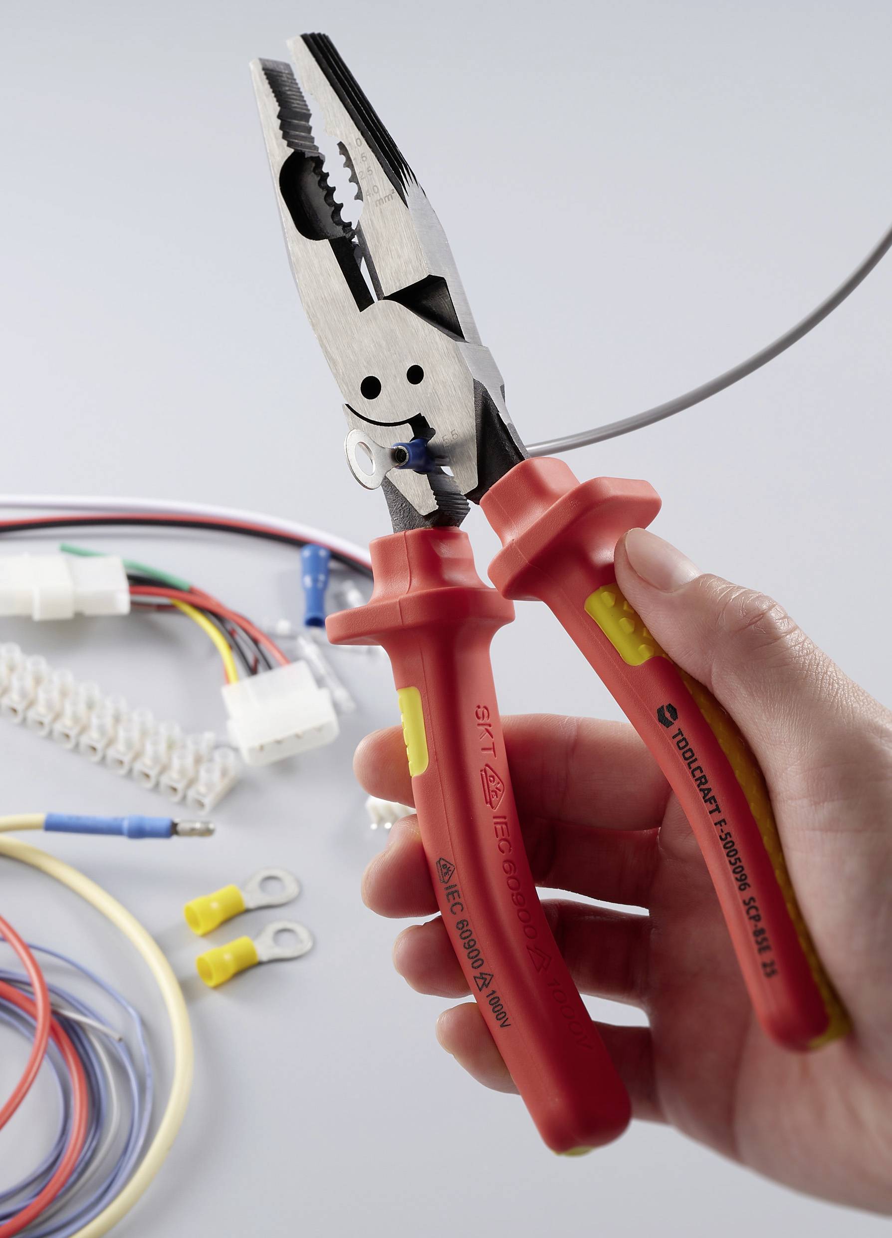 Eine Hand hält isolierte Zangen mit roten Griffen und veranschaulicht deren Einsatz bei elektrischen Arbeiten; verschiedene Kabel und Verbinder befinden sich im Hintergrund.