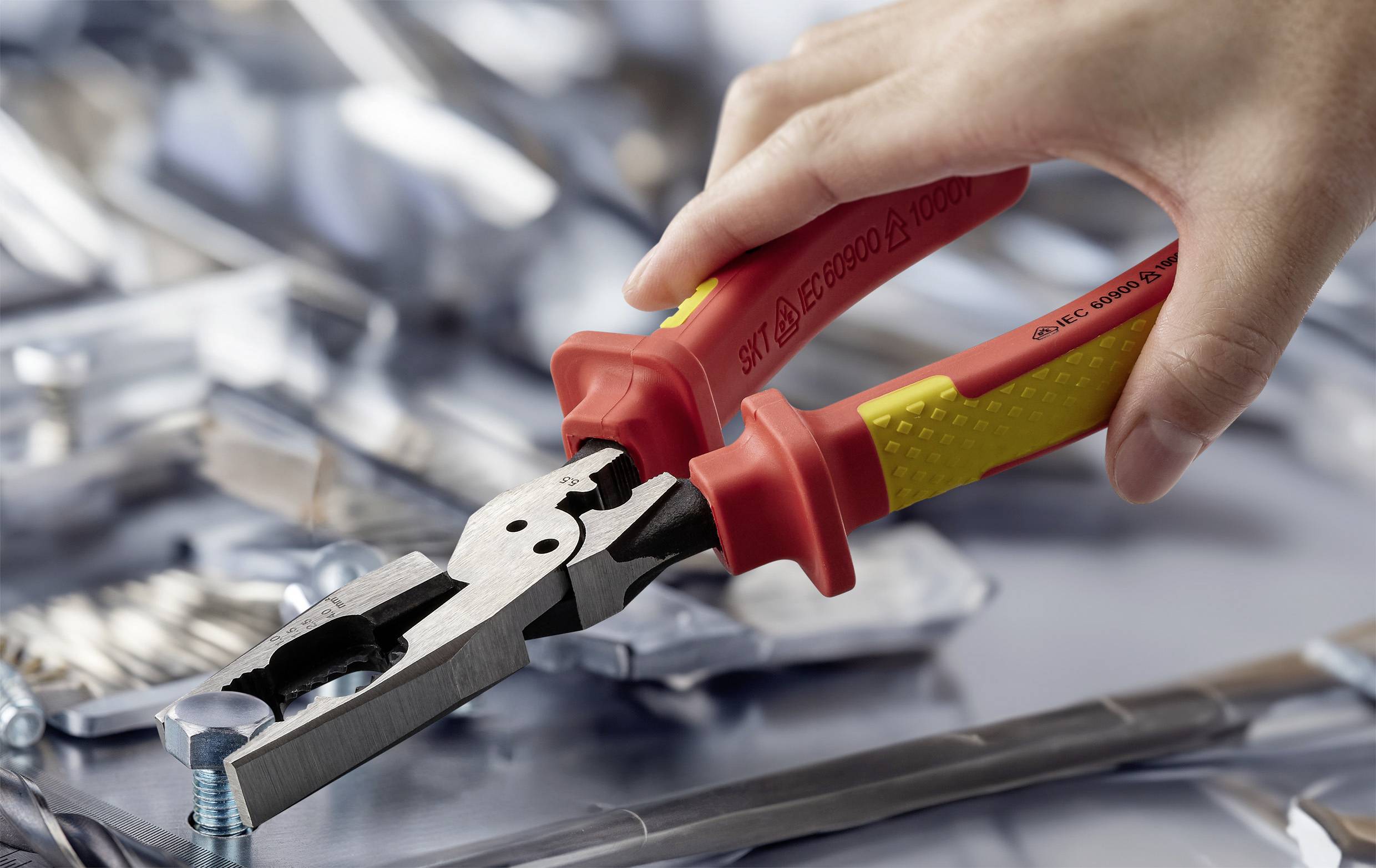 Eine Hand benutzt rote und gelbe Zangen, um eine Schraube auf einer reflektierenden Metalloberfläche festzuziehen, mit verstreuten Metallteilen im Hintergrund.