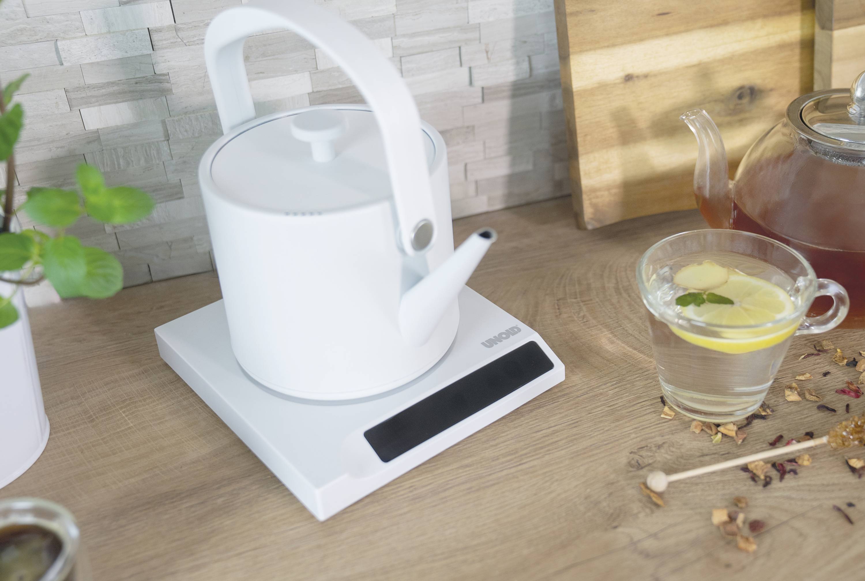 Ein weißer Wasserkocher steht auf einem Holzbrett in einer Küche. Daneben sind eine Tasse mit Zitronentee und ein Topf mit Tee zu sehen.