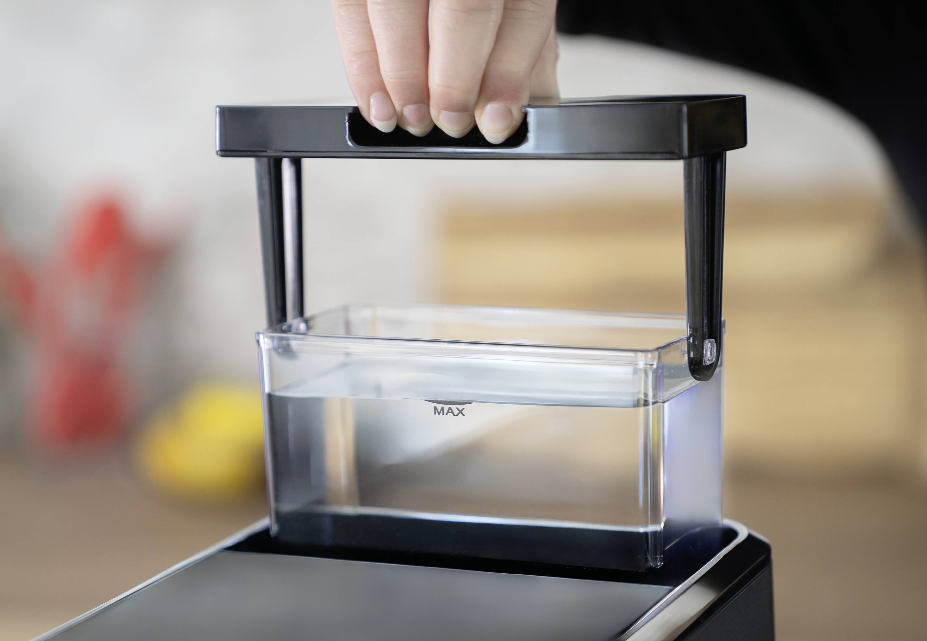 Eine Hand hebt einen Wassertank aus einer Maschine. Der Wassertank zeigt die 'MAX'-Markierung für die maximale Befüllung.