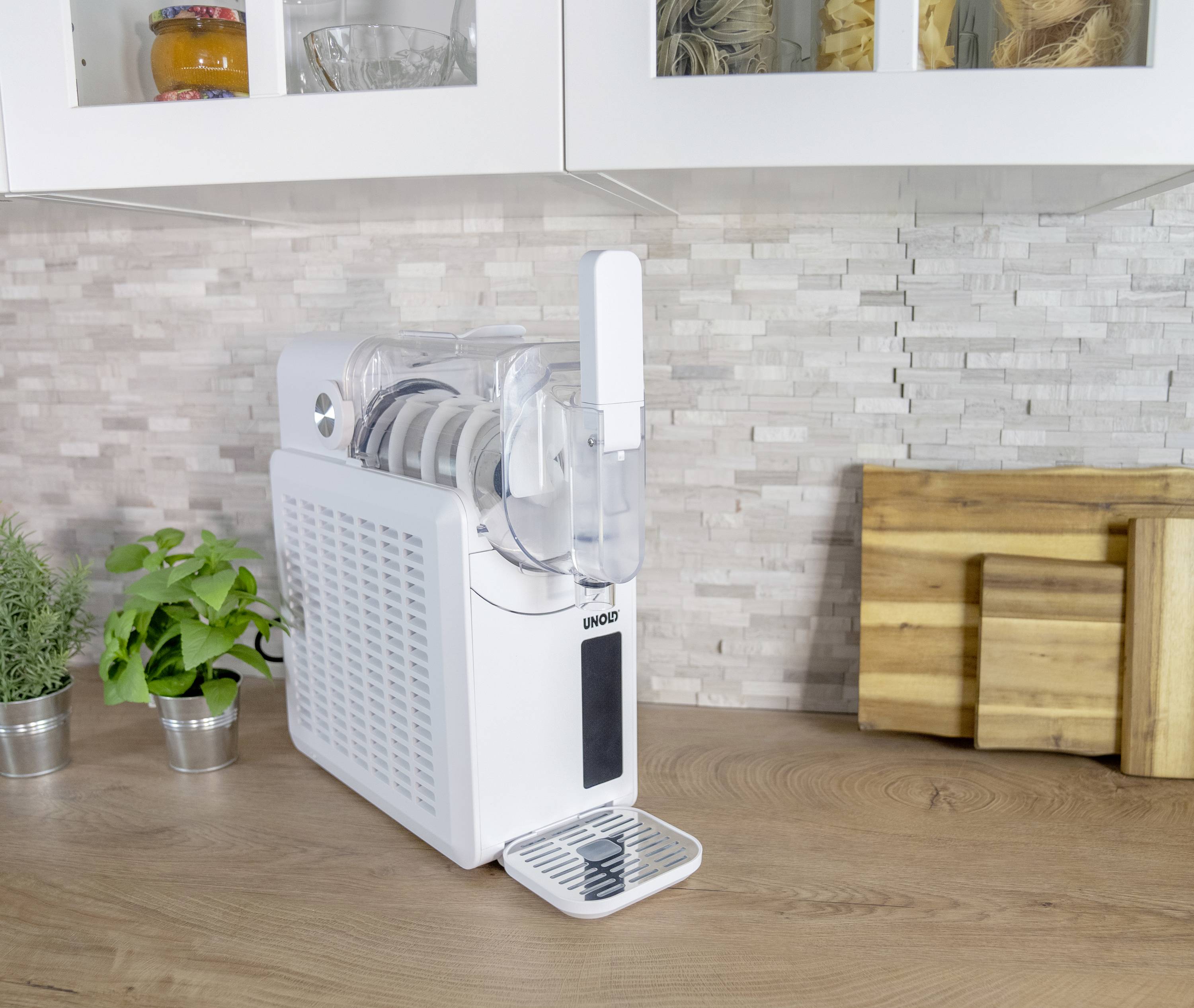 Ein weißer Wasserfilter auf einer Küchenarbeitsplatte. Im Hintergrund sind Holzschneidebretter, Pflanzen und Küchenutensilien zu sehen.