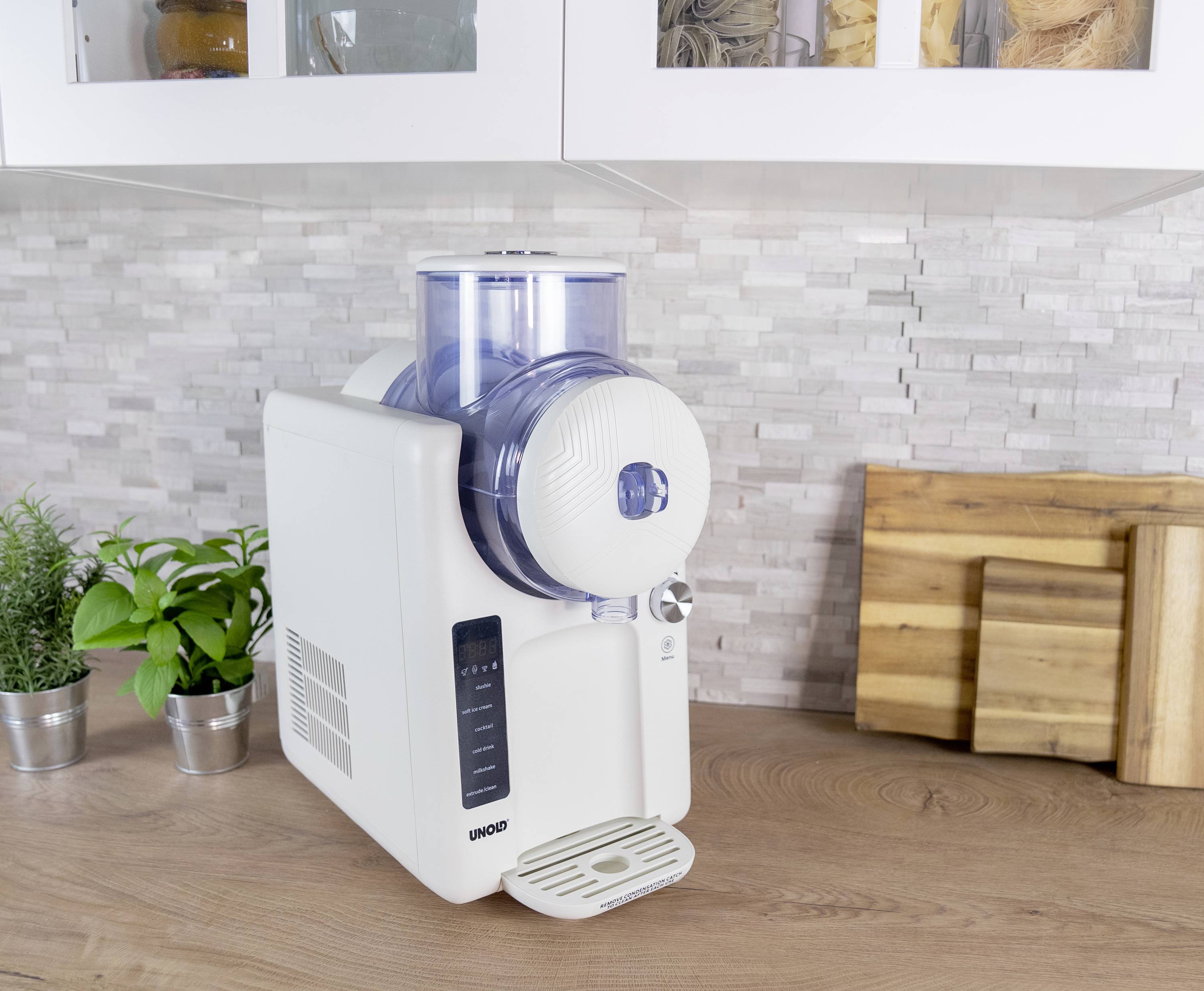 Eine weiße Küchenmaschine steht auf einer Holzarbeitsplatte. Daneben sind Pflanzen in Töpfen und Schneidebretter an der Wand.