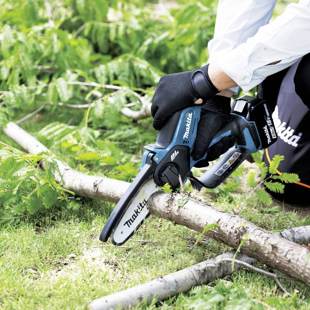 Eine Person mit Handschuhen verwendet eine tragbare elektrische Säge, um einen kleinen Ast zu schneiden, umgeben von grünem Laub, auf einer Rasenfläche.