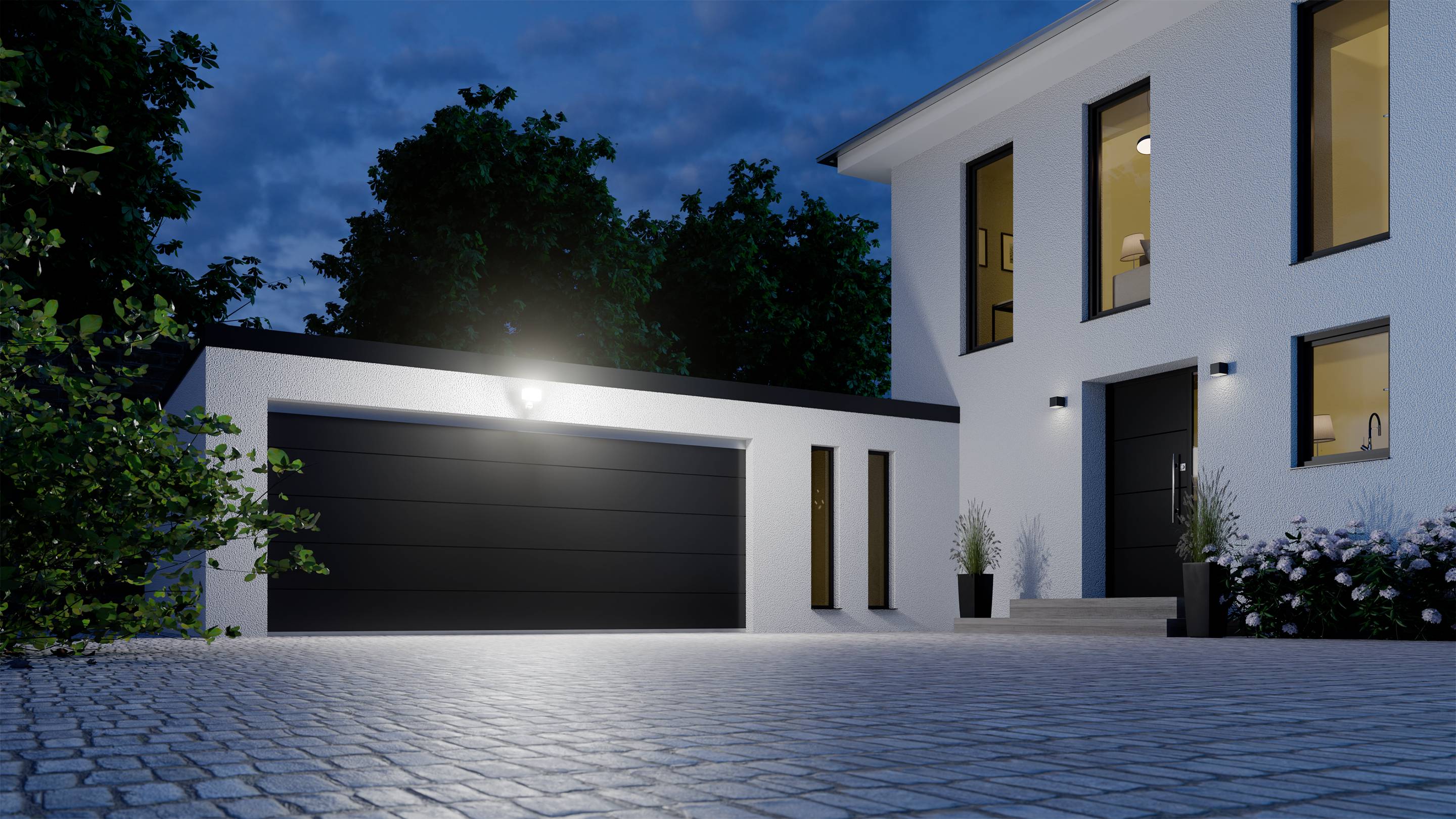 Modernes Haus bei Nacht, beleuchtet durch Außenlampen. Rechts ein zweistöckiges Gebäude, links eine Garage. Pflastersteine im Vordergrund.