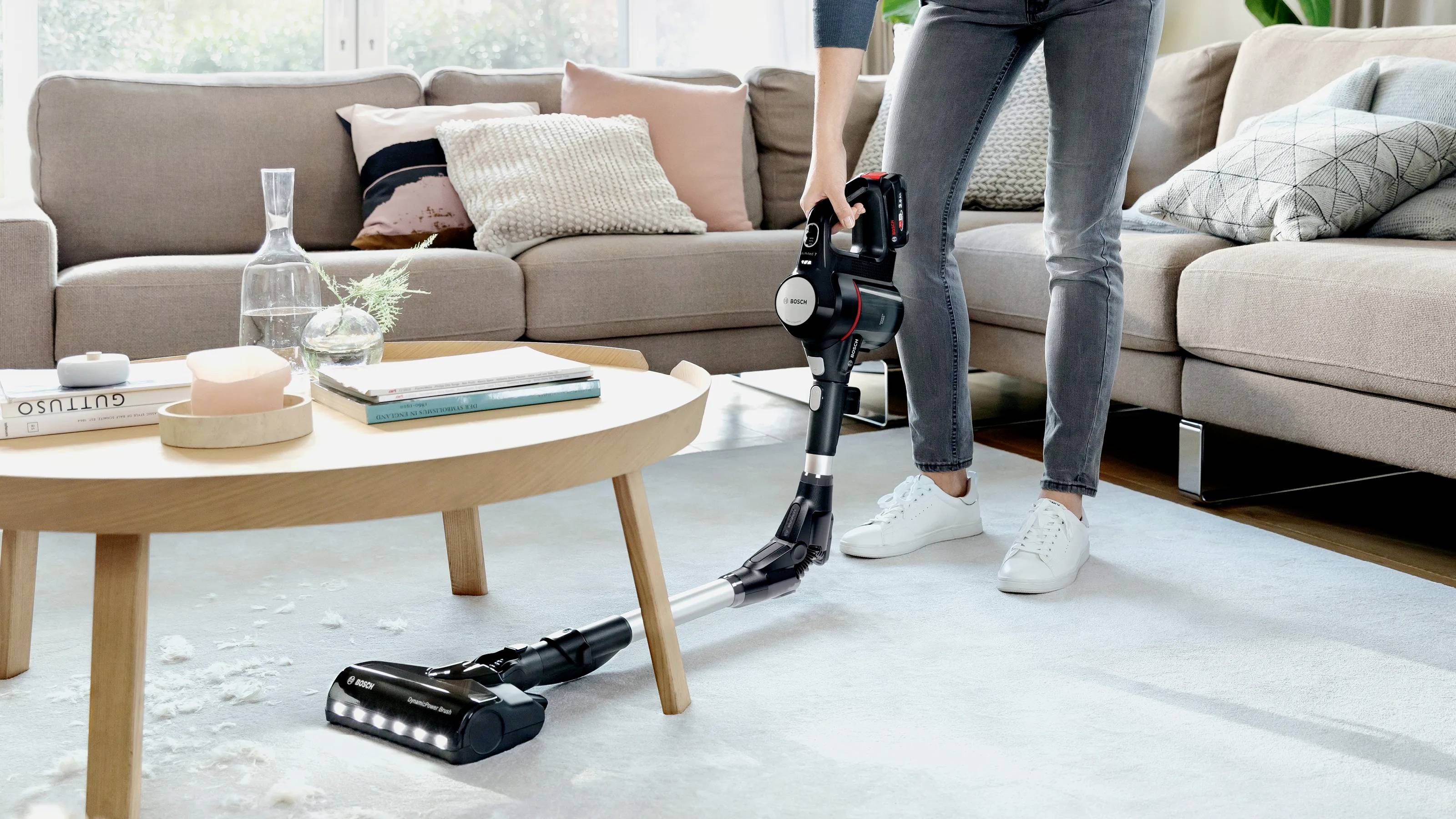 Eine Person saugt den Teppich in einem Wohnzimmer mit einem kabellosen Staubsauger. Im Hintergrund sind ein Sofa und ein Tisch zu sehen.