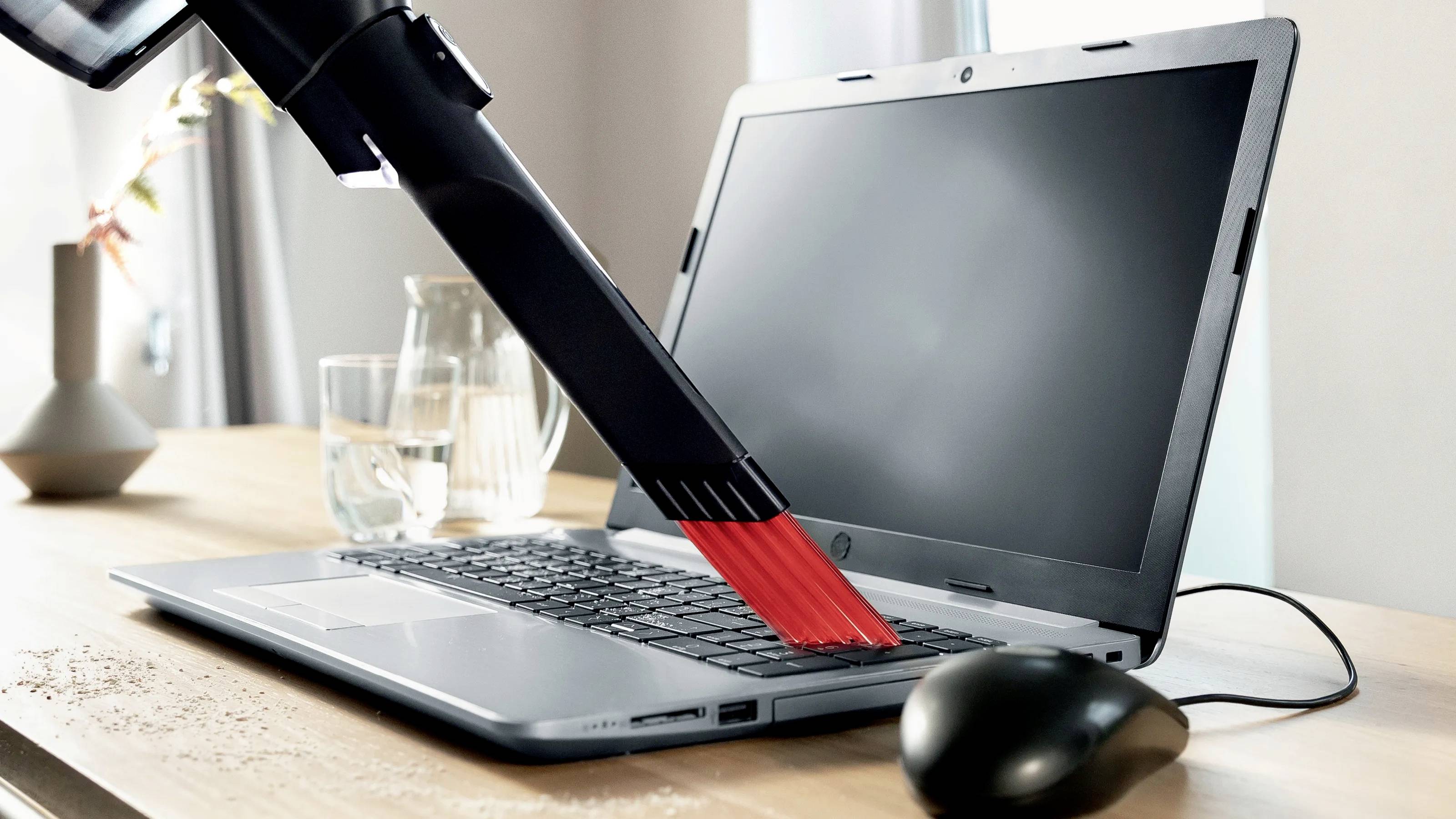 Ein Staubsauger saugt Krümel von einer Laptoptastatur auf. Im Hintergrund sind ein Glas Wasser und ein beleuchtetes Fenster zu sehen.