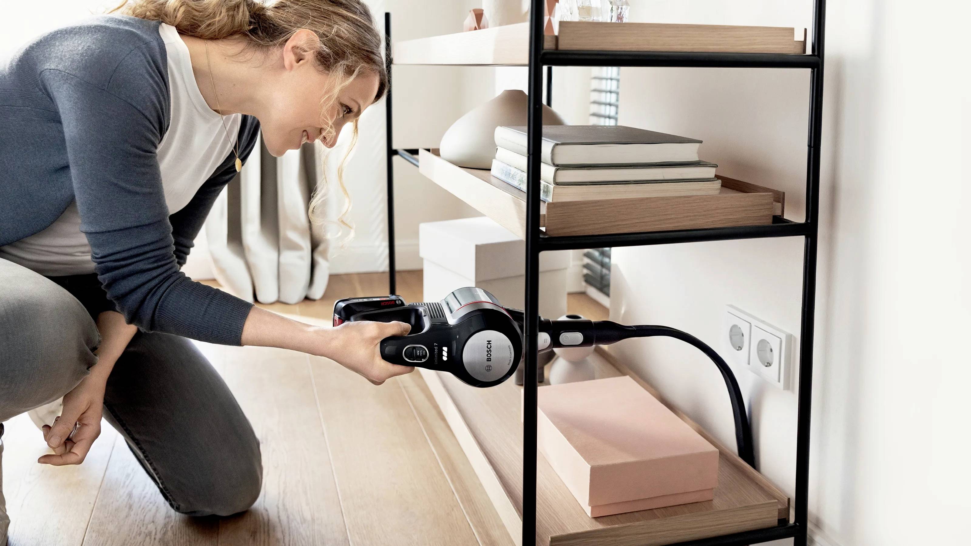 Eine Frau benutzt einen kabellosen Staubsauger, um den Boden unter einem Regal zu reinigen. Das Regal ist mit Büchern und Dekorationsgegenständen bestückt.