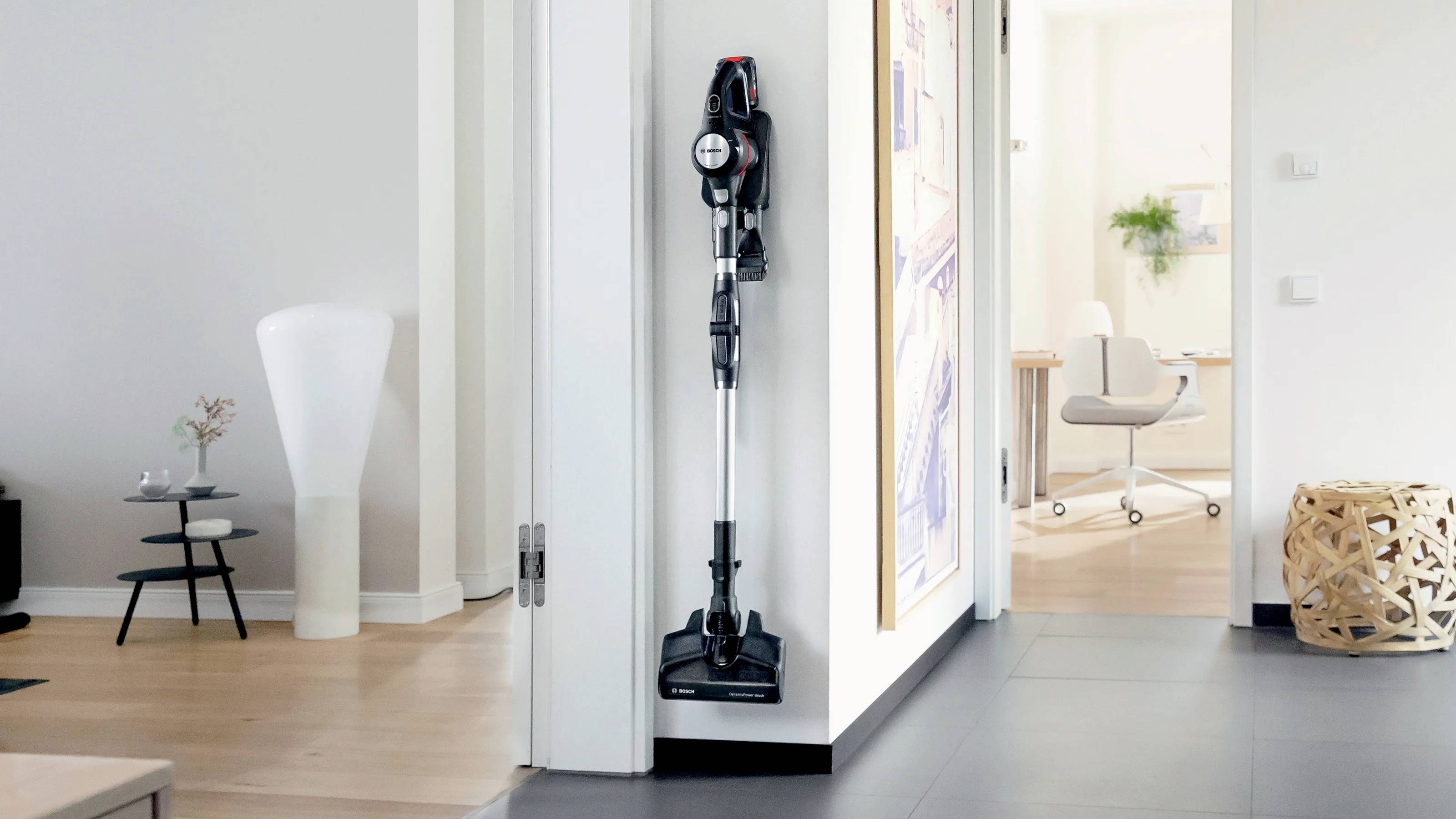 Ein kabelloser Staubsauger ist an der Wand in einem modernen Wohnzimmer befestigt. Nebenan ist ein offenes Büro zu sehen.