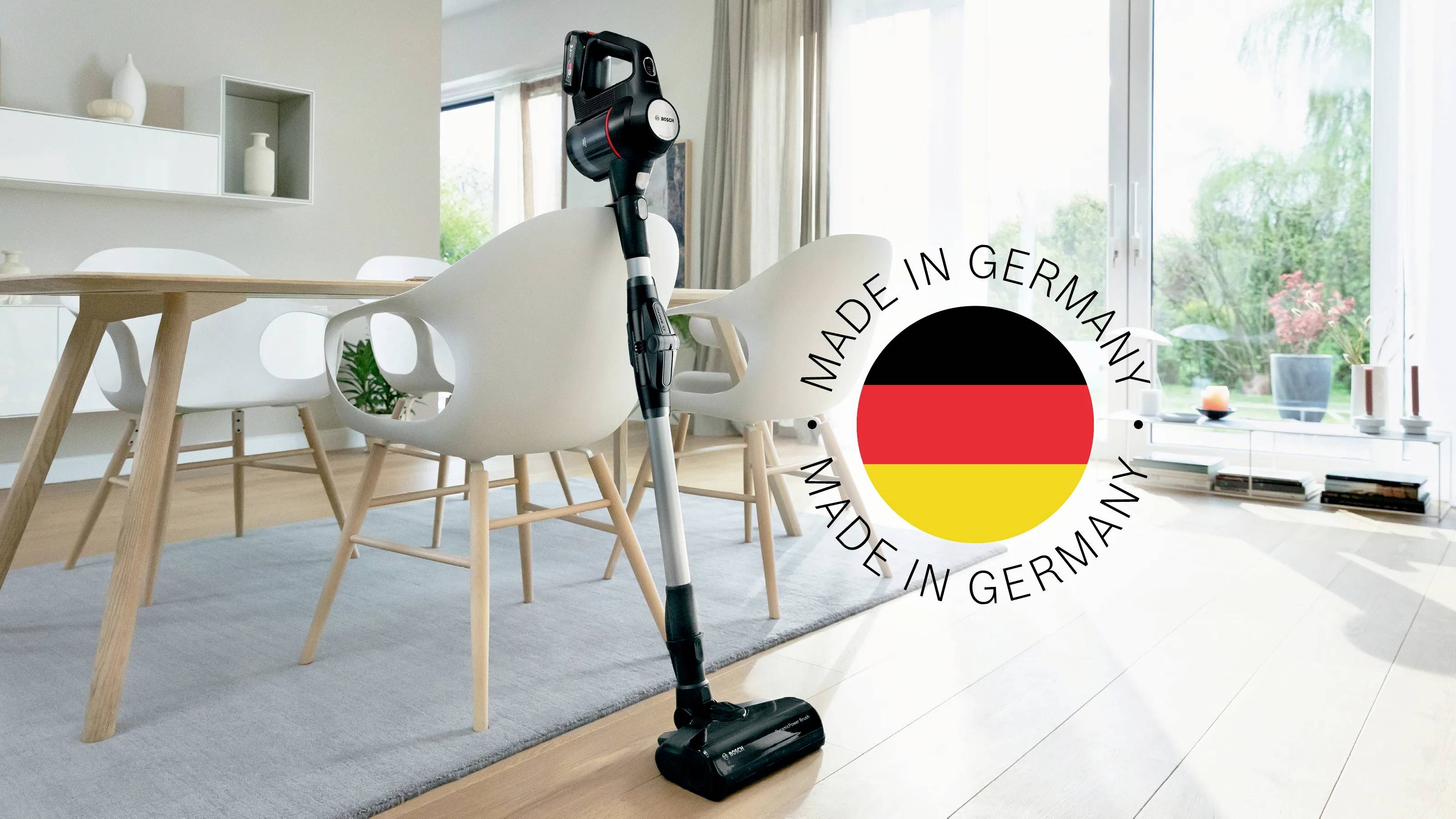 Ein kabelloser Staubsauger steht in einem modernen, hellen Wohnzimmer. Daneben ein Siegel mit 'Made in Germany' und der deutschen Flagge.