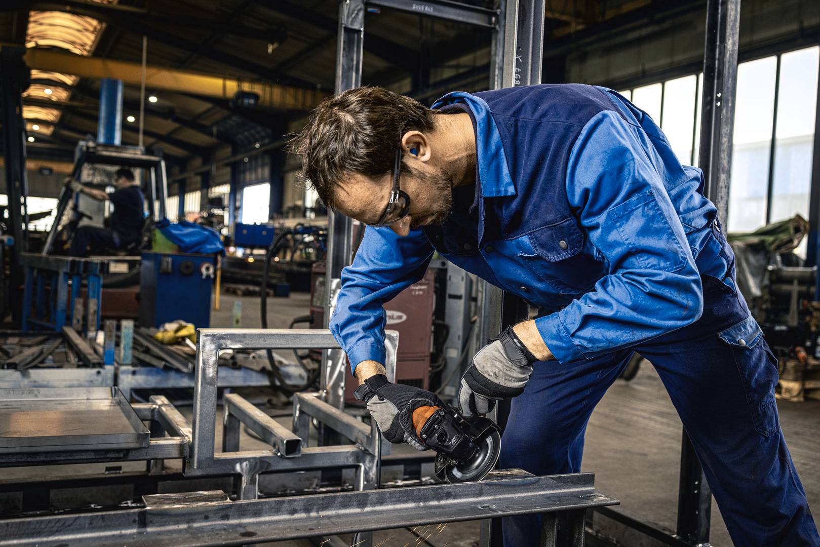 Ein Mann in Arbeitskleidung schleift Metall in einer Werkstatt. Im Hintergrund sind weitere Arbeiter und Ausrüstungen zu sehen.