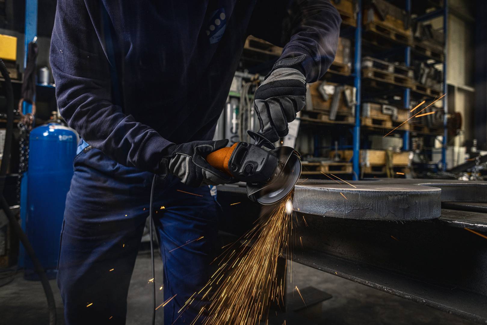 Eine Person schleift Metall mit einem Winkelschleifer in einer Werkstatt, Funken fliegen. Im Hintergrund sind Werkzeuge und Regale zu sehen.