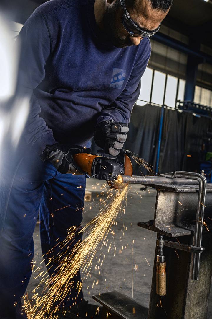 Ein Arbeiter in Schutzkleidung schleift ein Metallstück mit einem Winkelschleifer. Funken sprühen, deutet auf Metallverarbeitung hin.