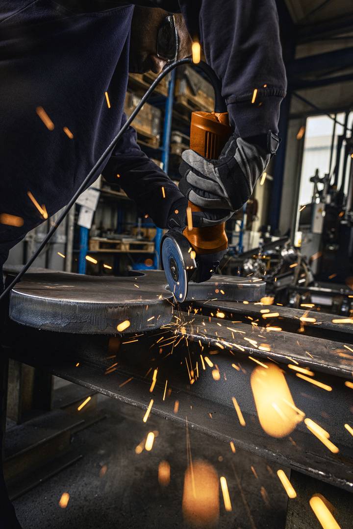 Person schleift Metall in Werkstatt, Funken fliegen. Schutzkleidung trägt zur Sicherheit bei. Industrielle Umgebung sichtbar im Hintergrund.