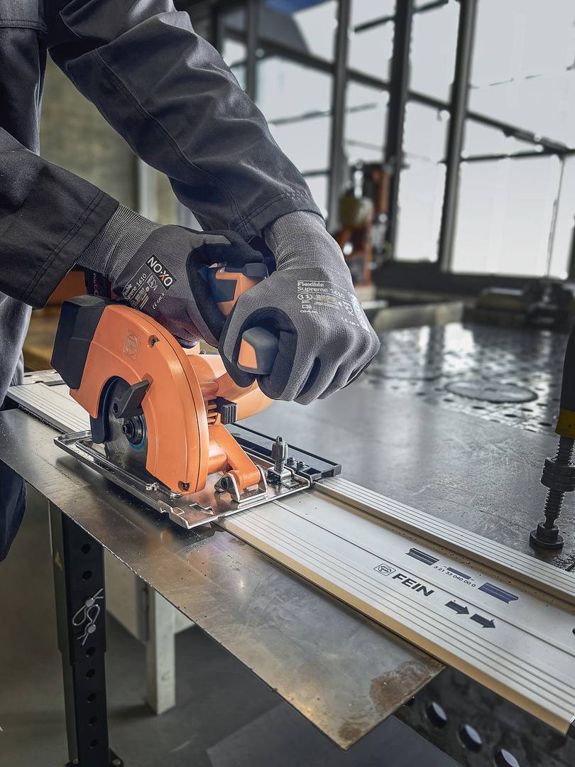 Eine Person schneidet mit einer orangefarbenen Handkreissäge entlang einer Schiene ein Metallstück. Schutzkleidung und Handschuhe sind zu sehen.