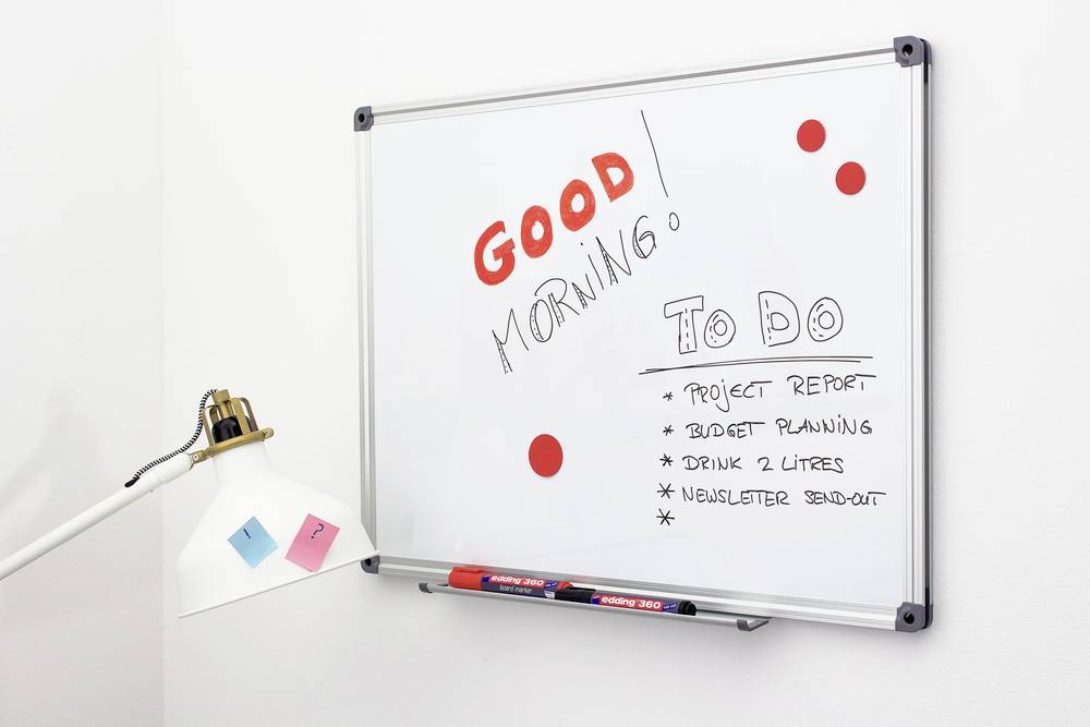 Whiteboard mit der Aufschrift 'Good morning! To do: Project report, Budget planning, Drink 2 liters, Newsletter send-out', daneben eine Lampe.