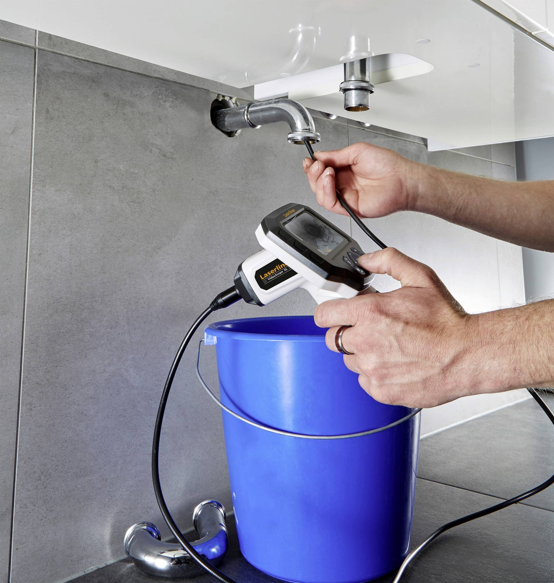 Eine Person verwendet ein Inspektionsgerät, um das Innere eines Abflussrohrs unter einem Waschbecken zu überprüfen. Ein blauer Eimer steht darunter.