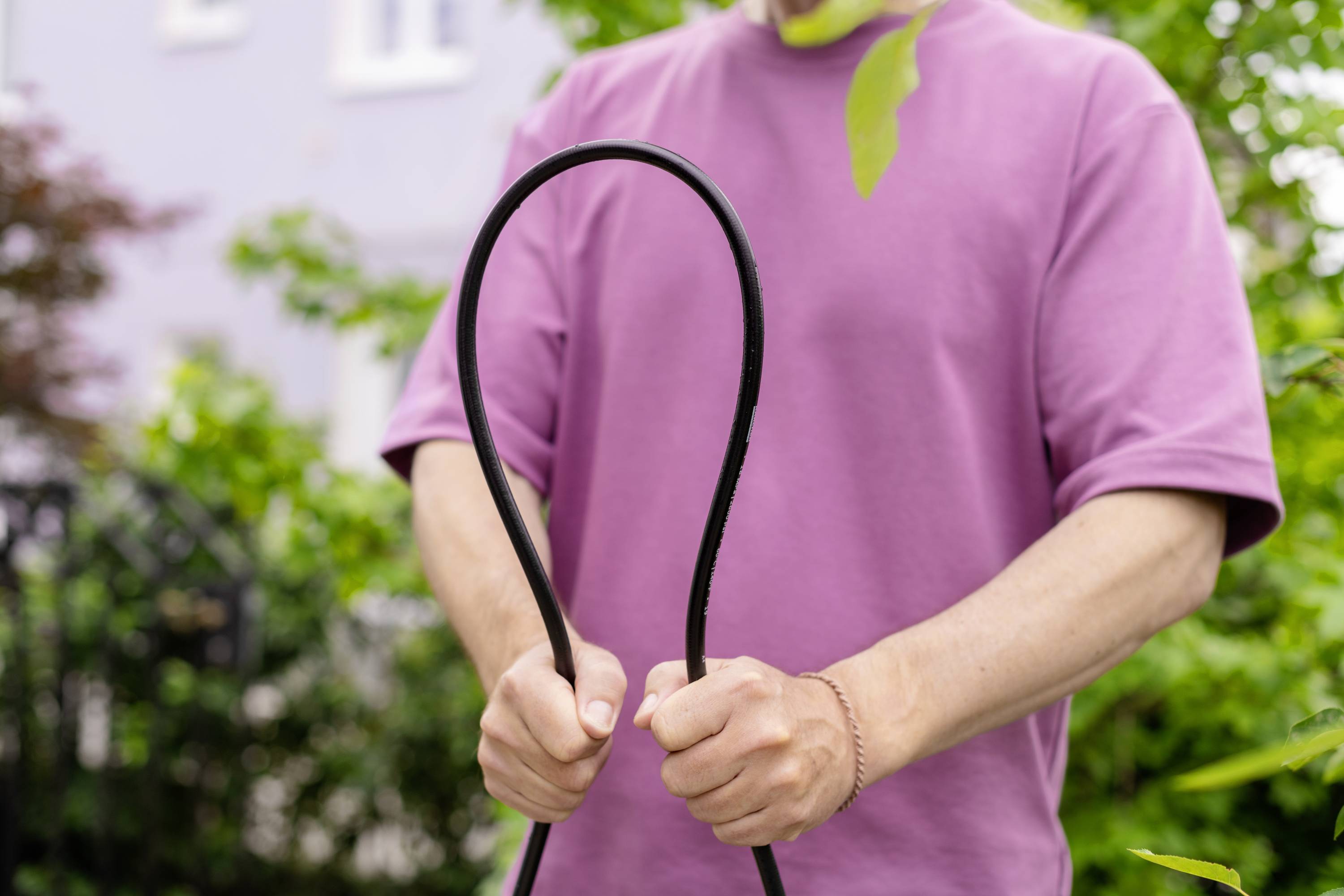 Person im rosa T-Shirt hält einen schwarzen Schlauch, der zu einer Schlaufe geformt ist, im Freien. Im Hintergrund sind Pflanzen zu sehen.