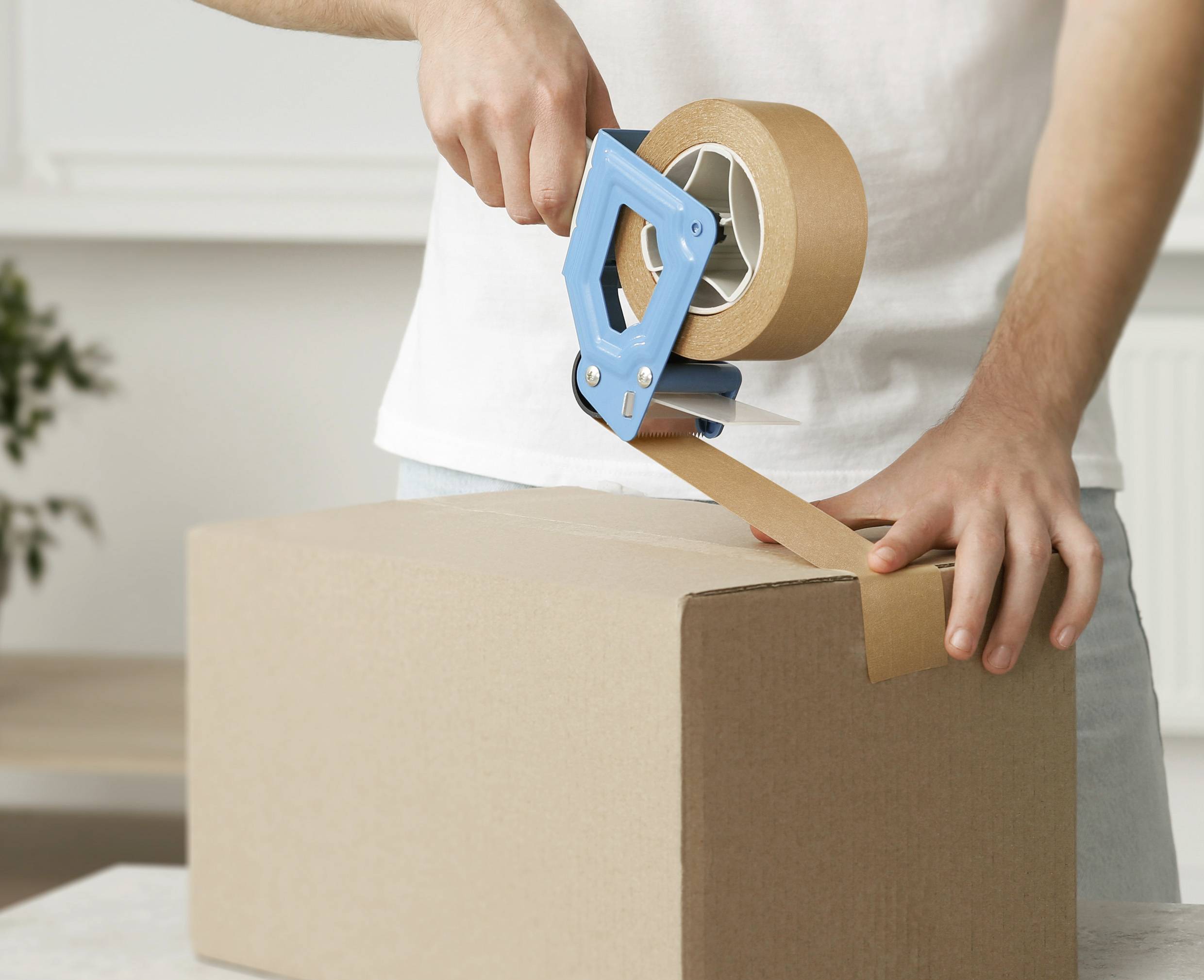 Eine Person verschließt einen Karton mit braunem Klebeband und einem blauen Abroller.