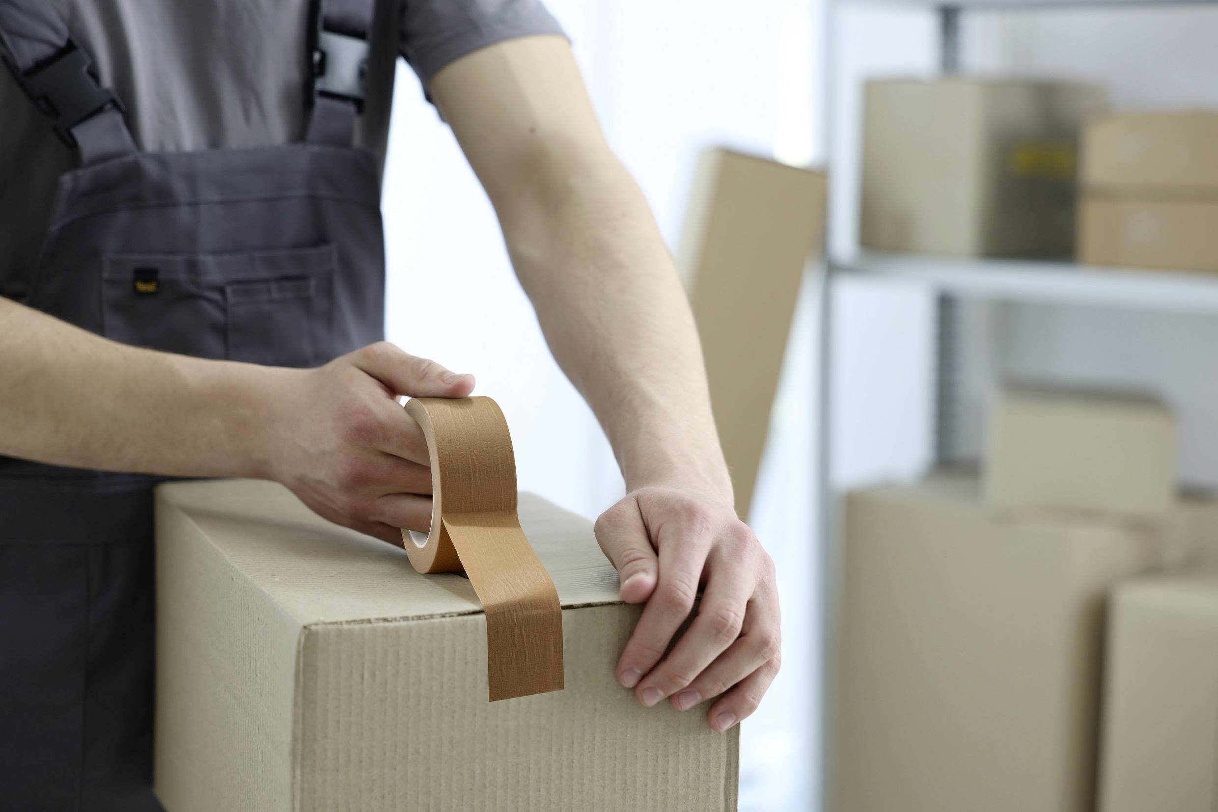 Eine Person in Arbeitskleidung klebt ein Paket zu. Im Hintergrund sind weitere Kartons und Regale zu sehen, möglicherweise in einem Lager.