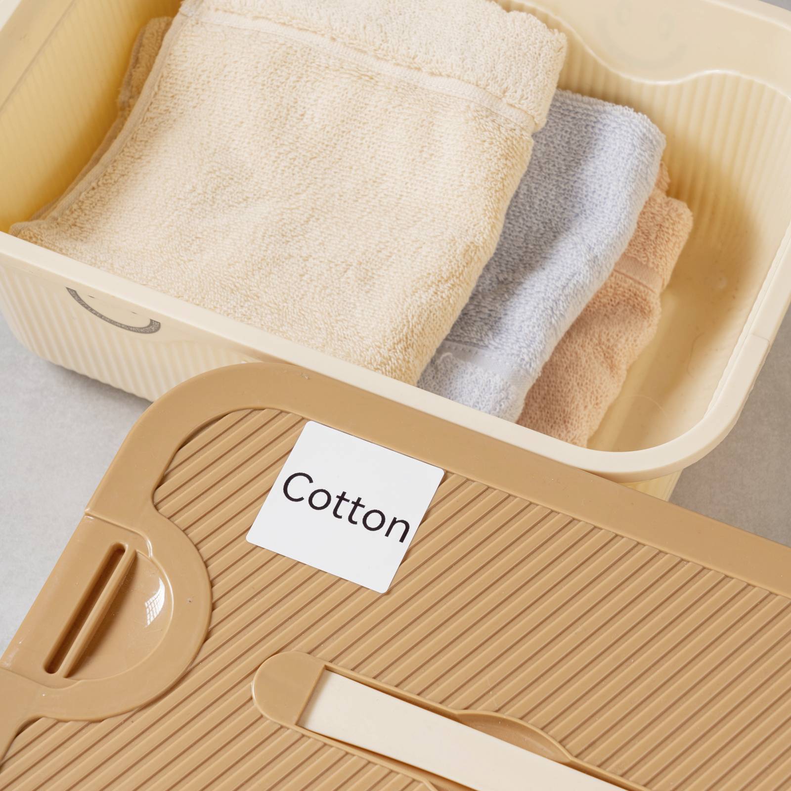 Ein Korb mit gefalteten Handtüchern, darunter ein beigefarbenes, ein graues und ein weiteres in Beige. Daneben liegt der Deckel mit dem Etikett 'Cotton'.