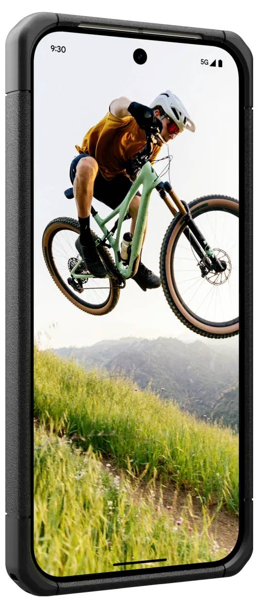 Ein Mountainbiker in orangem Trikot und Helm springt mit seinem Fahrrad in der Luft über eine grüne Wiese, im Hintergrund Berge.