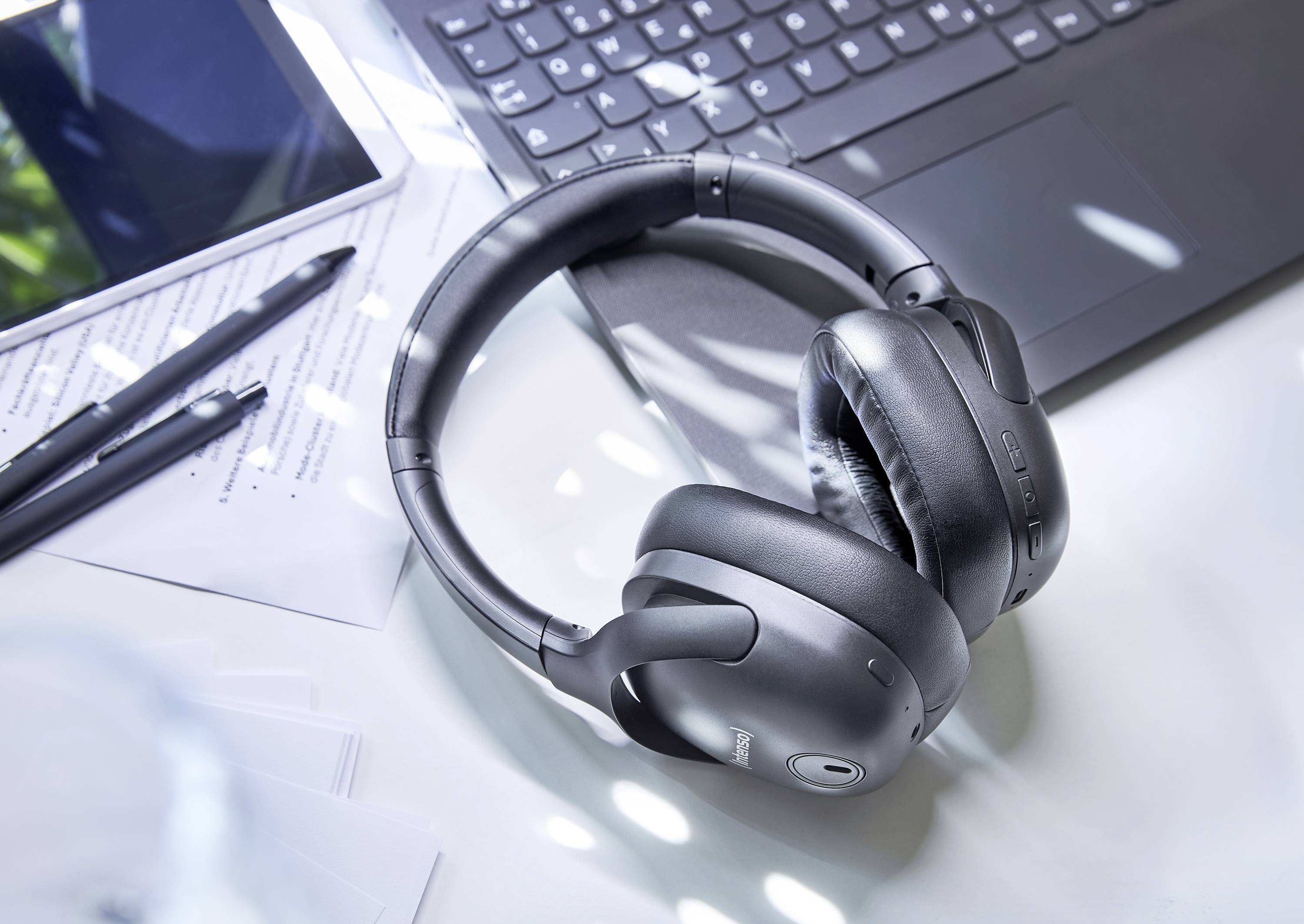 Kopfhörer liegen auf einem weißen Schreibtisch neben einem Laptop, einem Tablet und Notizunterlagen mit Stift.