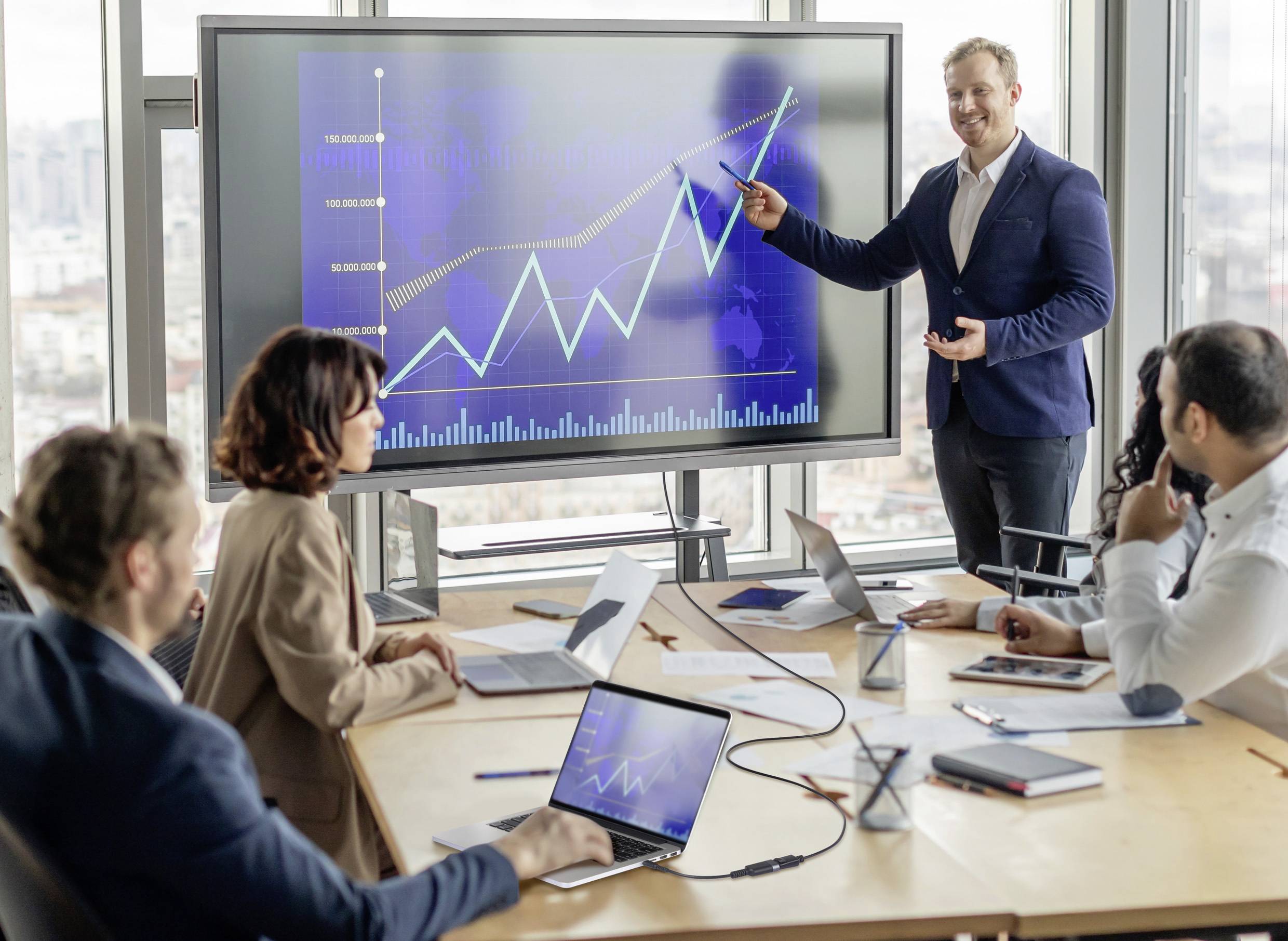 Ein Geschäftsmann präsentiert ein Diagramm mit steigenden Trends auf einem Bildschirm, während vier Personen aufmerksam in einem Konferenzraum zuhören.