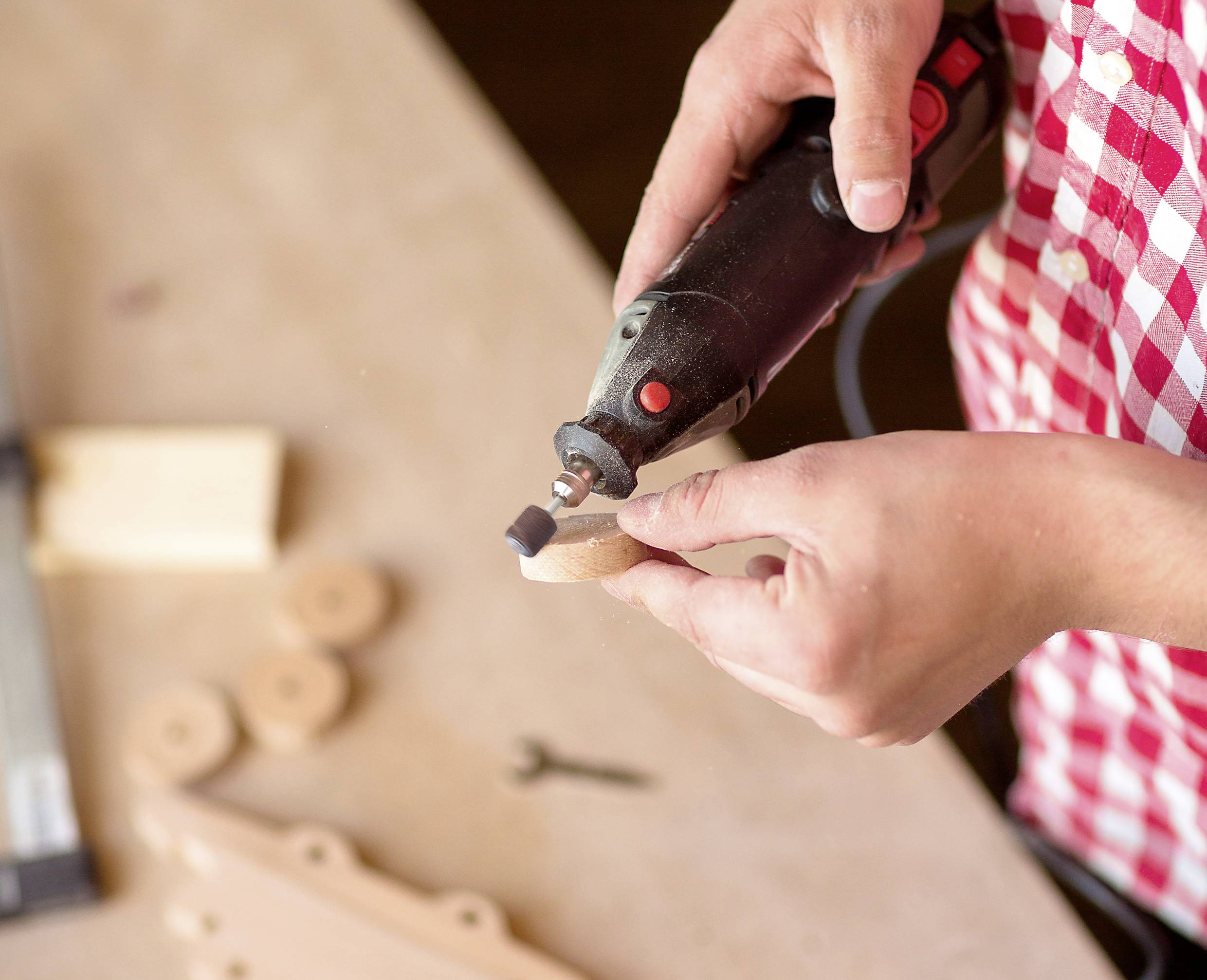 Eine Person in einem rot-weiß karierten Hemd benutzt ein Rotationswerkzeug, um ein Holzobjekt auf einer Werkbank zu schleifen, wobei zusätzliche Teile und ein Werkzeug in der Nähe liegen.