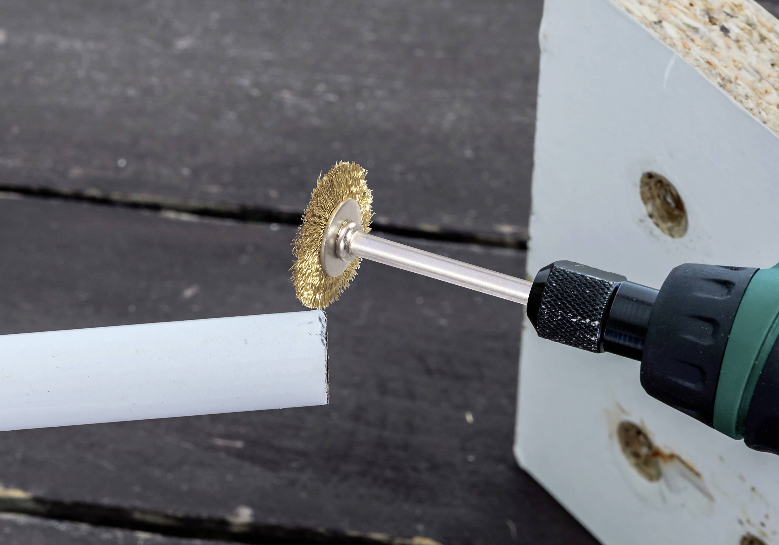 Eine Rotationsmaschine mit einer Drahtbürsten-Aufsatz reinigt oder poliert das Ende eines weißen zylindrischen Objekts, höchstwahrscheinlich ein Rohr, in der Nähe eines Holzblocks.