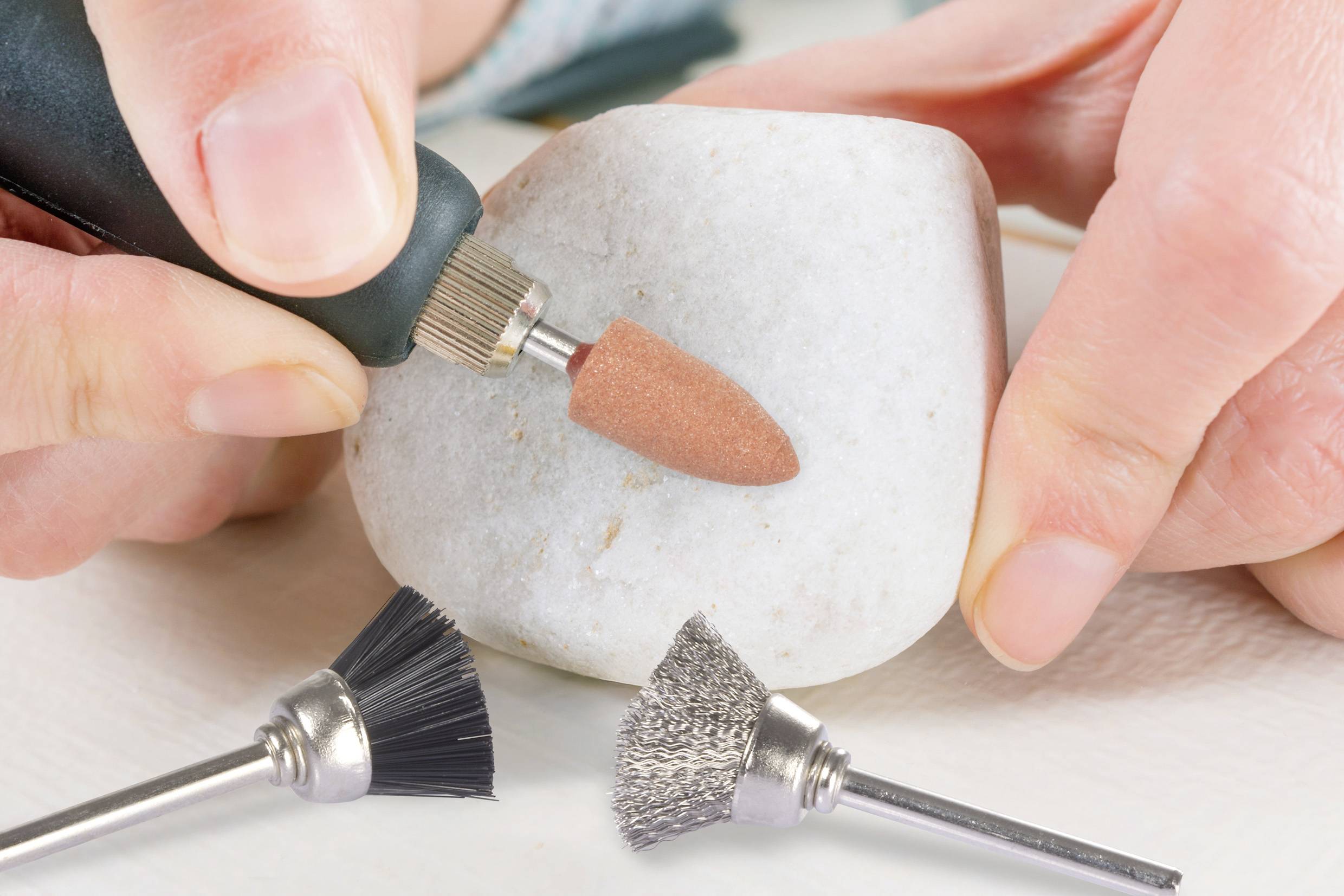 Eine Person verwendet ein Rotationswerkzeug, um einen weißen Stein zu formen. Zwei verschiedene Bürstenaufsätze liegen im Vordergrund und deuten auf eine Bastel- oder Heimwerkeraktivität hin.