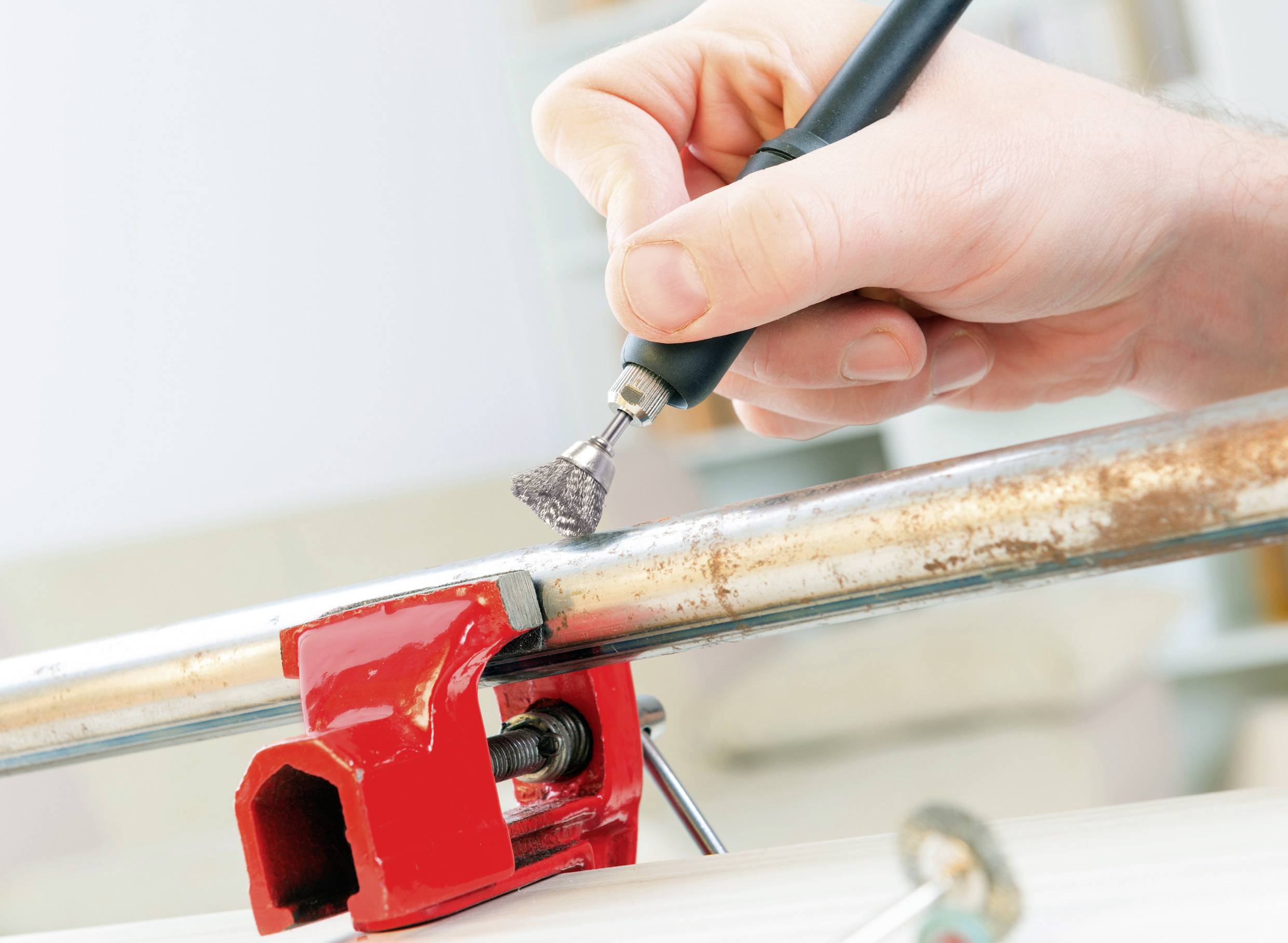 Eine Hand verwendet ein Rotationswerkzeug mit Bürstenaufsatz, um Rost von einem Metallrohr zu entfernen, das in einem roten Schraubstock befestigt ist.