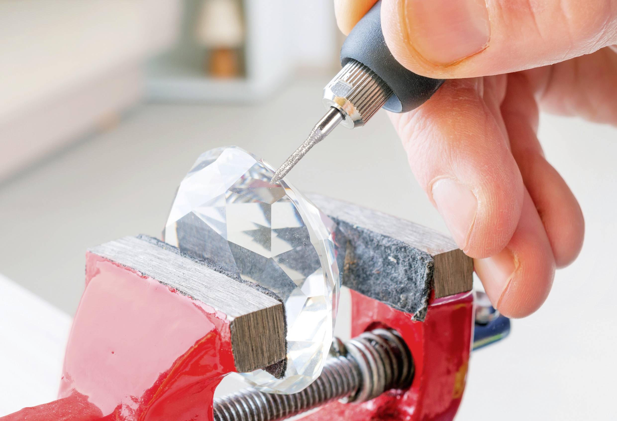 Eine Hand benutzt ein Rotationswerkzeug, um einen großen Kristall zu schneiden, der in einem roten Schraubstock gesichert ist, was auf eine präzise Edelsteingravur oder -formgebung hindeutet.