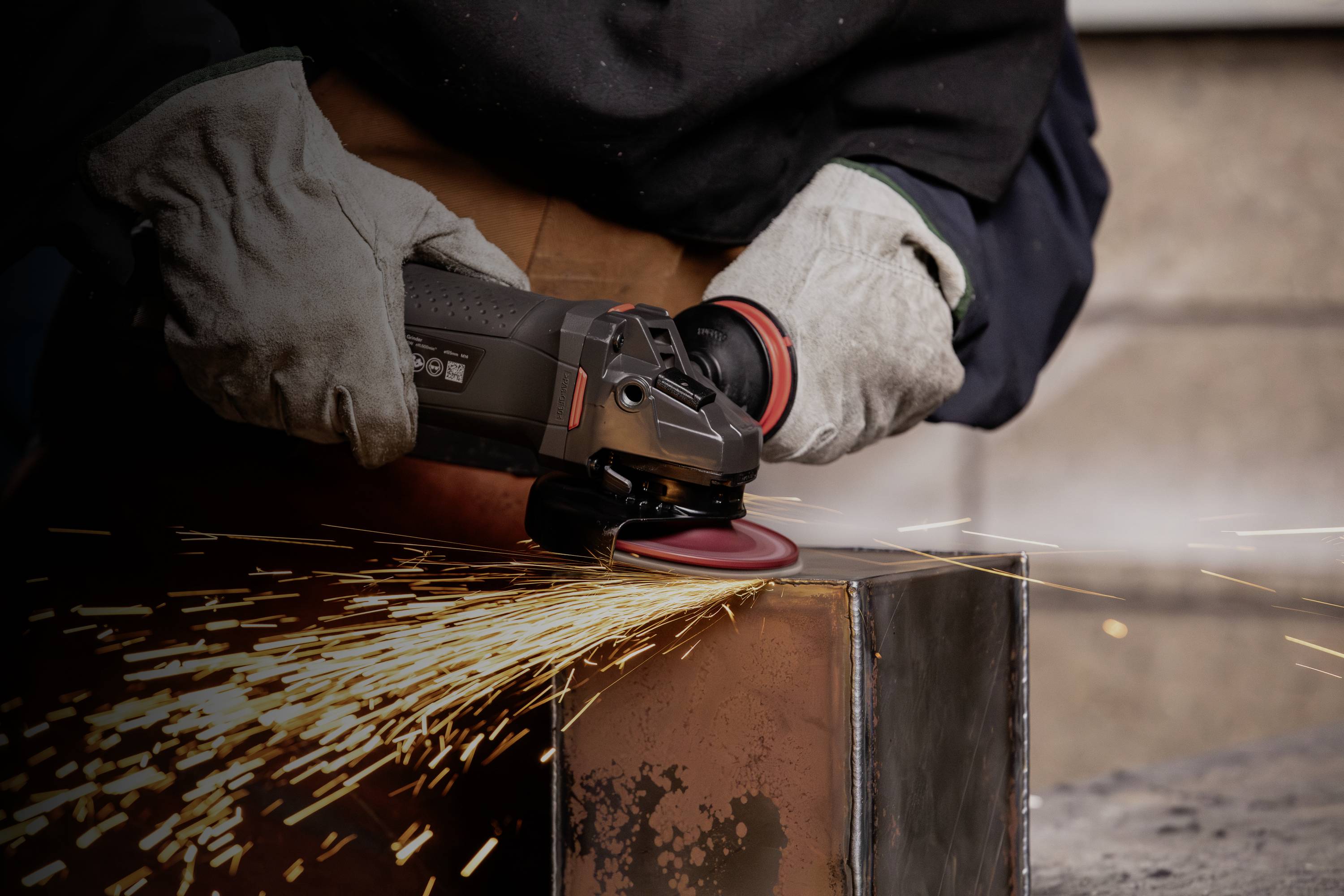 Eine Person schleift mit einem Winkelschleifer Metall. Funken fliegen von der Metalloberfläche. Die Person trägt Schutzhandschuhe.