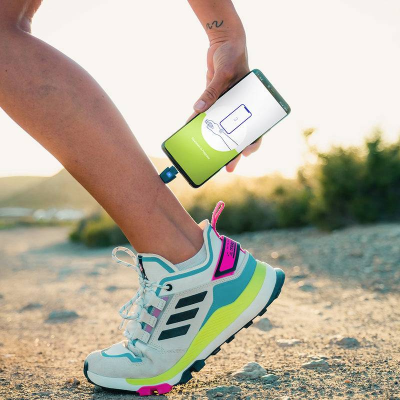 Eine Person trägt sportliche Schuhe und verwendet ein Smartphone, das mit einem kleinen Gerät verbunden ist, um wahrscheinlich Schritte oder Bewegung zu messen.