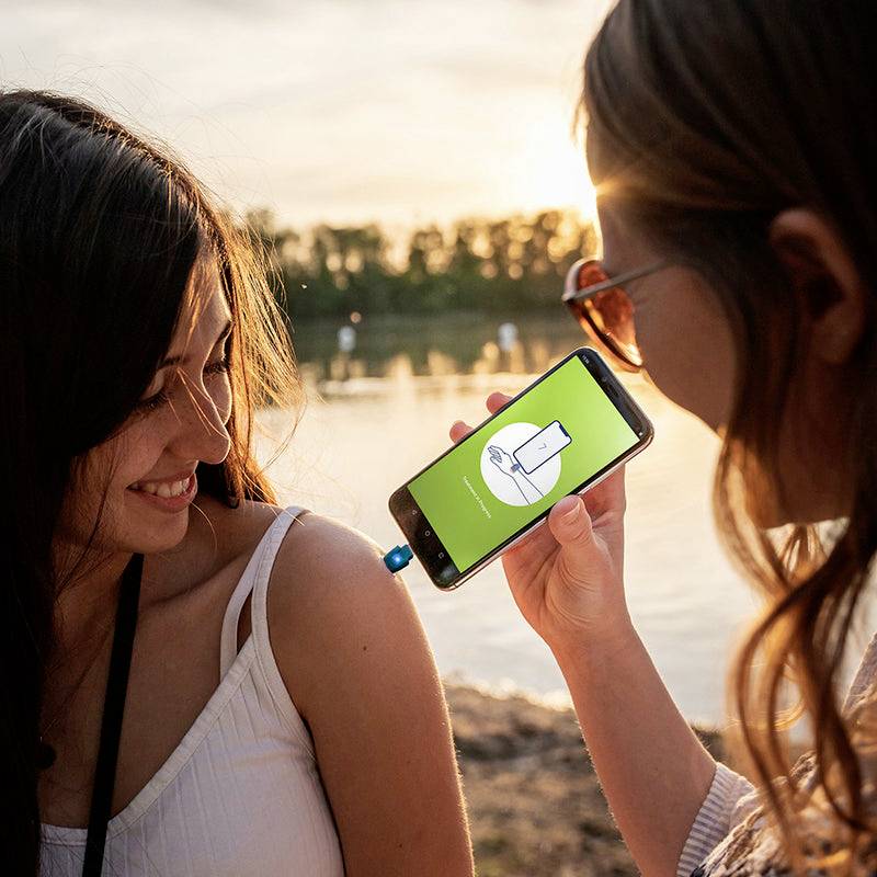 Zwei Personen stehen im Freien am Wasser. Eine Person zeigt der anderen eine Handy-App mit Bild eines Hörgeräts. Sie lächeln.