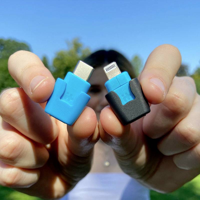 Person hält zwei Adapter, einen USB-C-Adapter in Blau und einen Lightning-Adapter in Schwarz, vor einem verschwommenen Hintergrund.