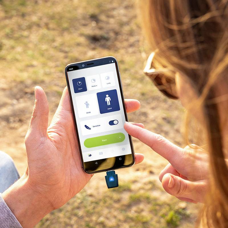 Person hält Smartphone mit Gesundheits-App; zeigt Symbole für Herz, Lunge und Schritte zählen. Gerät mit blauem Stecker verbunden.