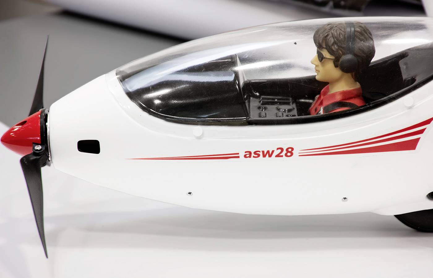 Ein Modellflugzeug in Nahaufnahme, mit einem detailreichen Cockpit und einem Pilotenmodell. Beschriftung 'asw28' und aufgedruckte rote Linien.