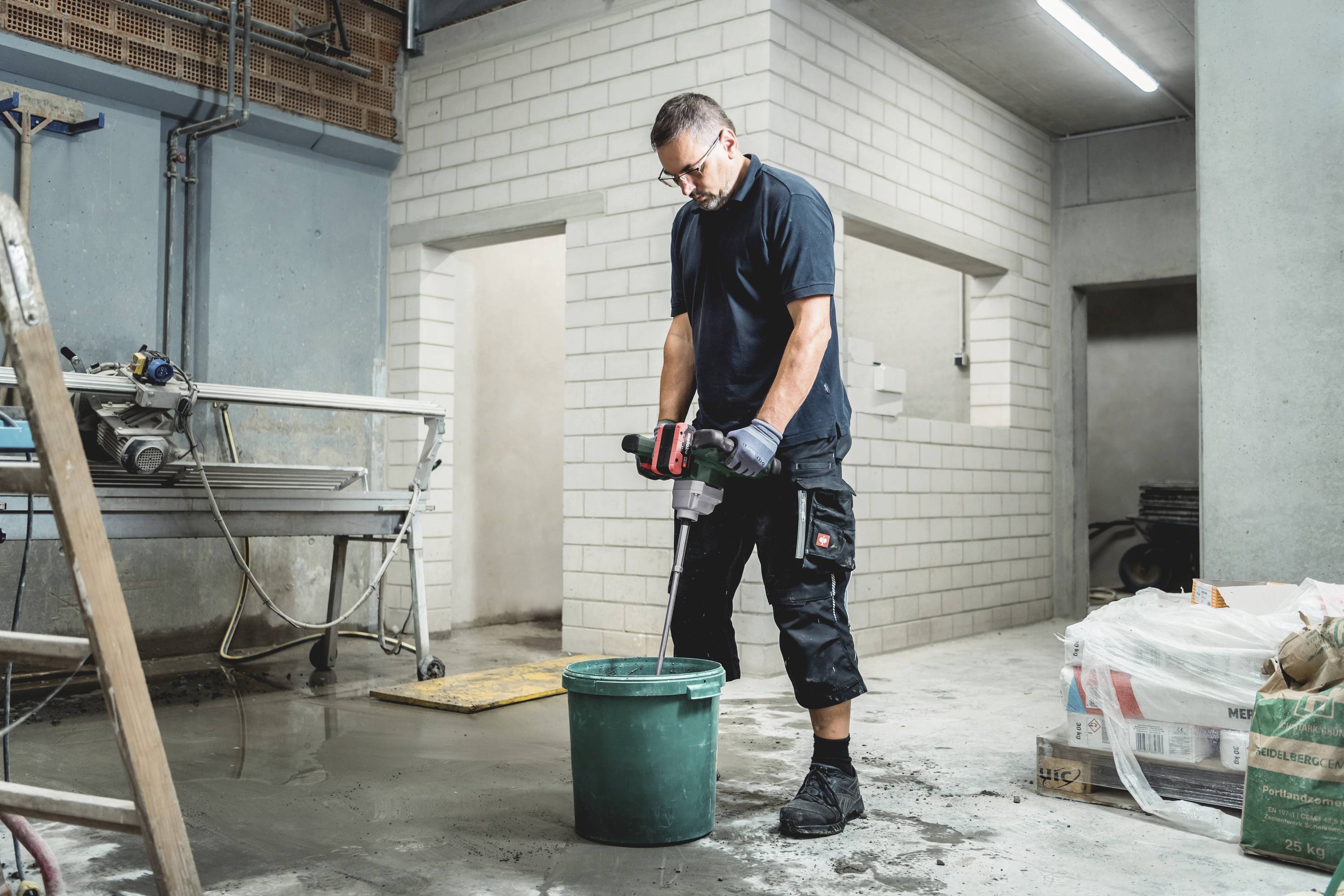 Ein Mann in Arbeitskleidung mischt mit einem elektrischen Rührwerk Material in einem Eimer auf einer Baustelle.