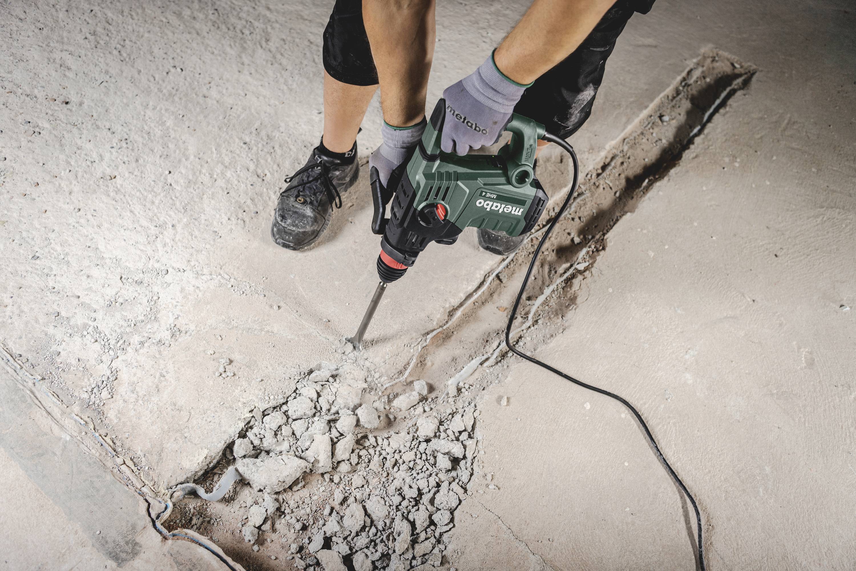 Person, die mit einem grünen Bohrhammer einen Betonboden aufbricht. Der Boden ist teilweise aufgebrochen, und Trümmer liegen herum.