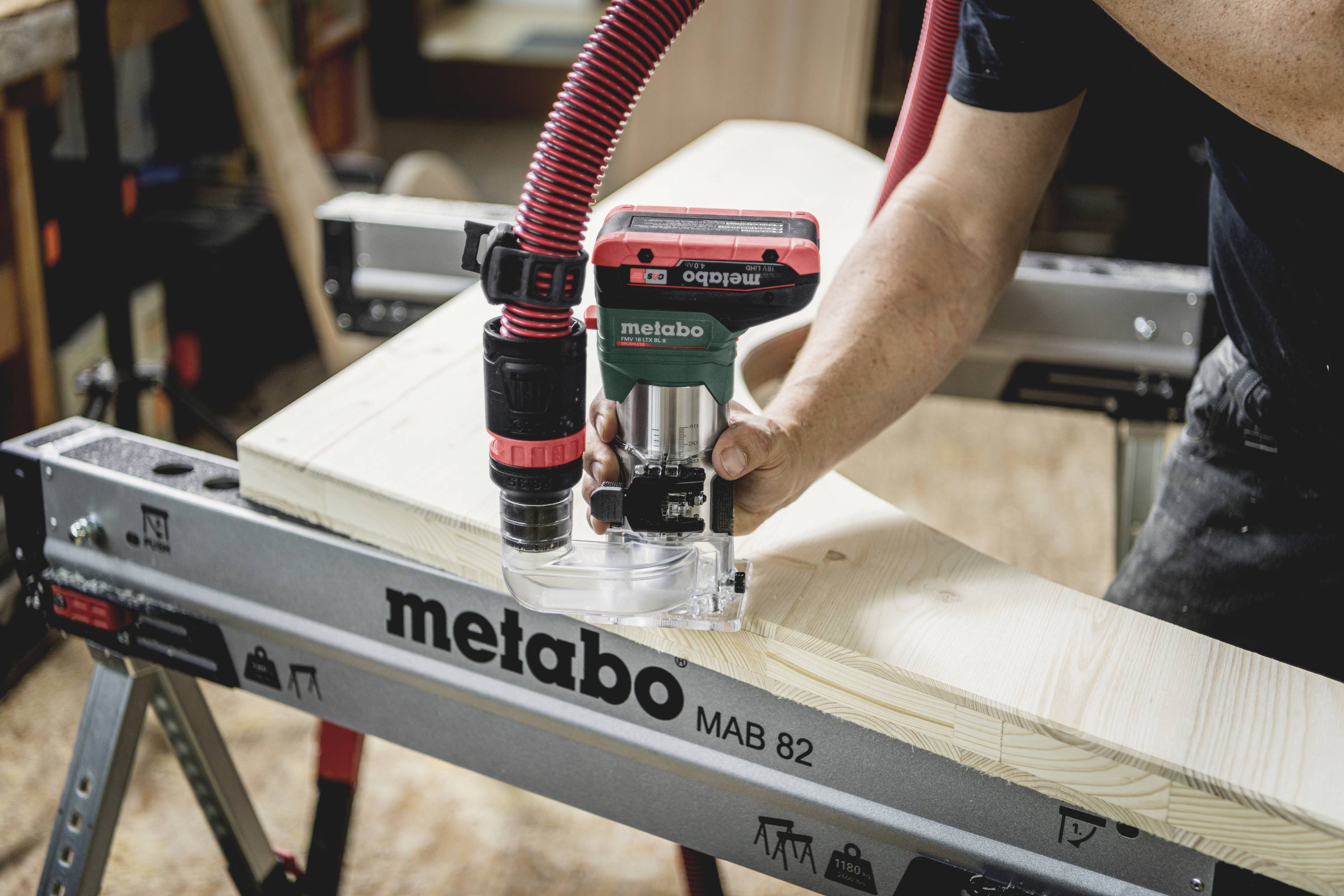 Eine Person arbeitet mit einer Handoberfräse auf einem hellen Holzstück, die Fräse ist auf einem Metabo-Werkzeugtisch befestigt.